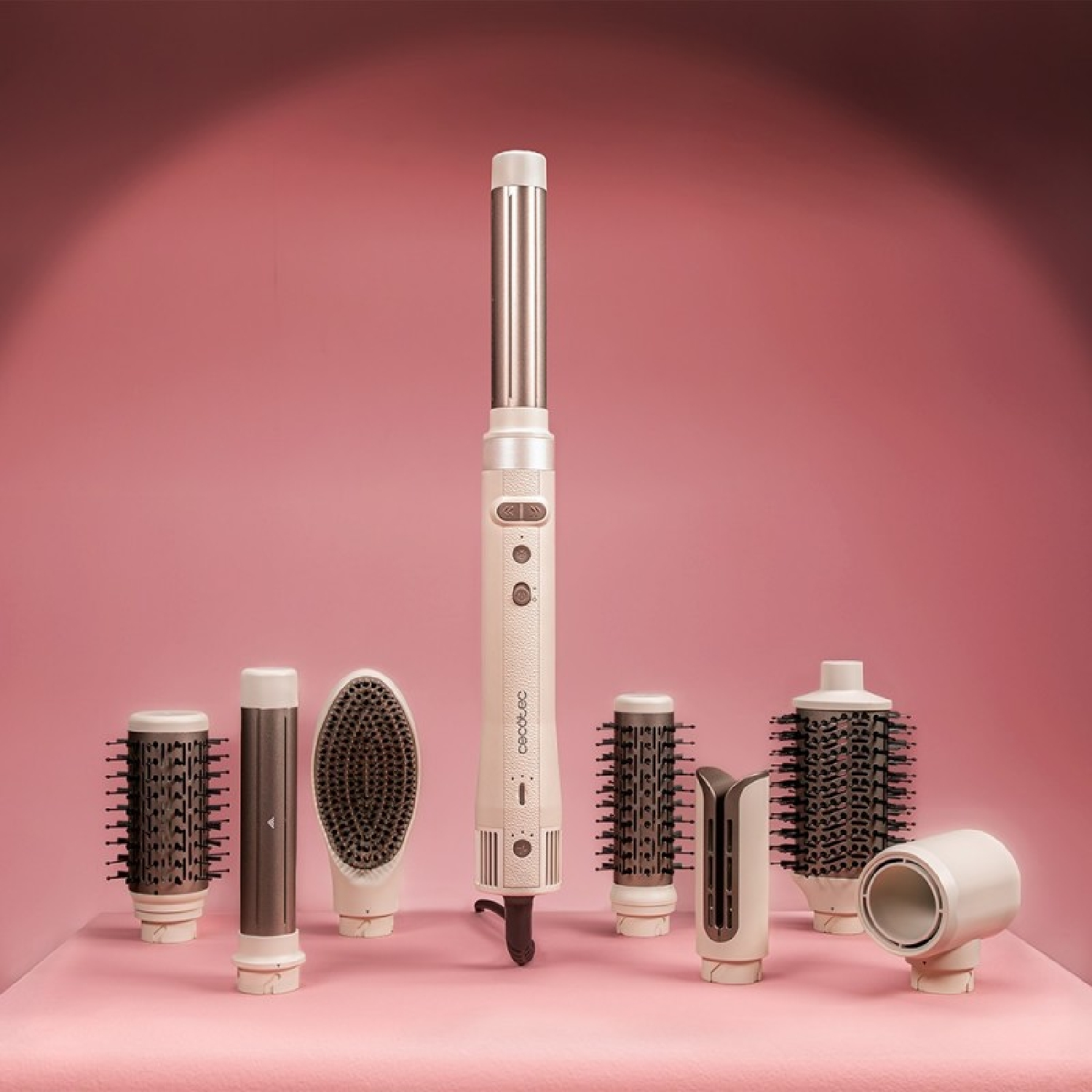 Un set di strumenti per lo styling dei capelli, tra cui un ferro arricciacapelli e vari attacchi per spazzole, disposti su una superficie rosa.