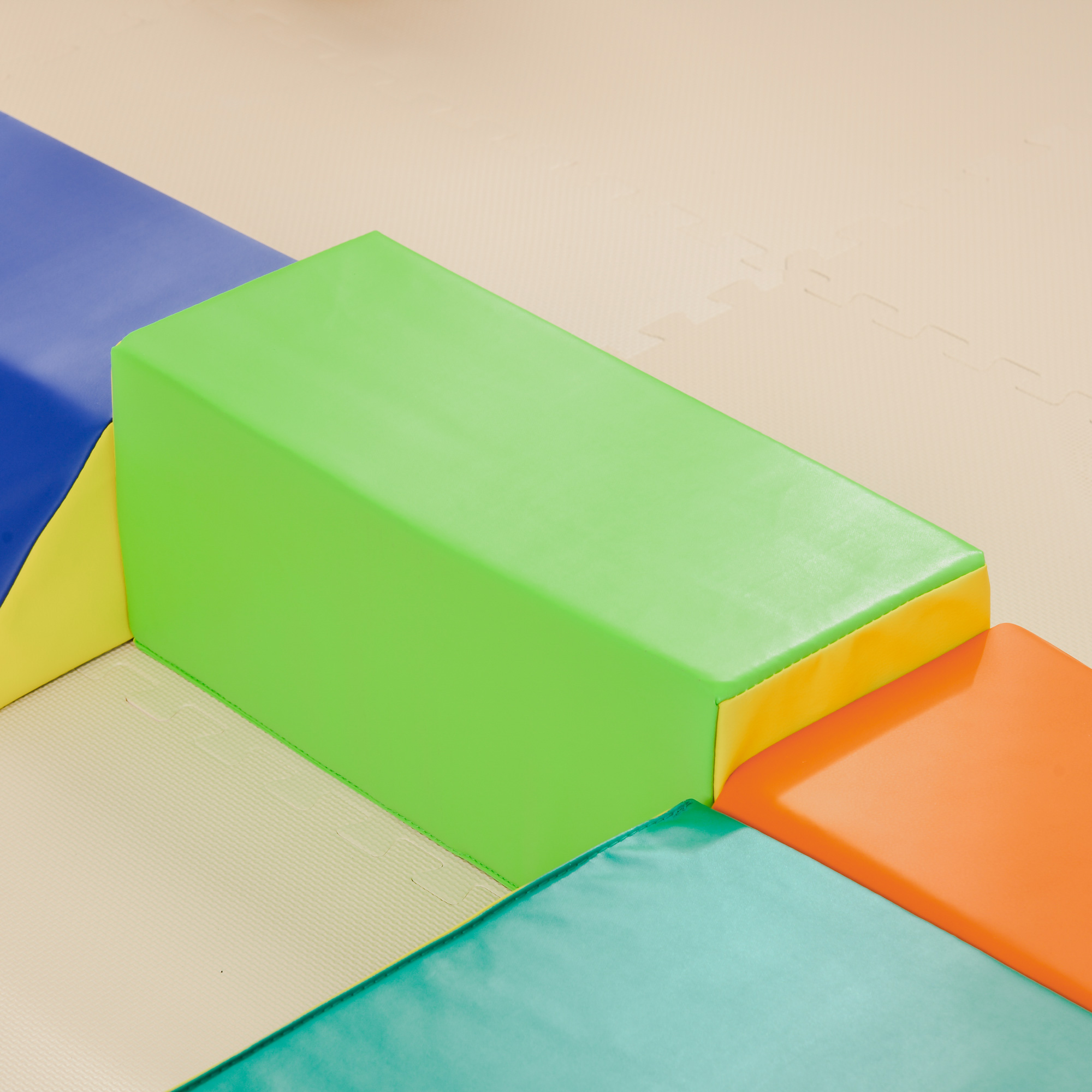 Una struttura di gioco colorata con blocchi verdi, arancioni, blu e gialli su un pavimento imbottito beige.