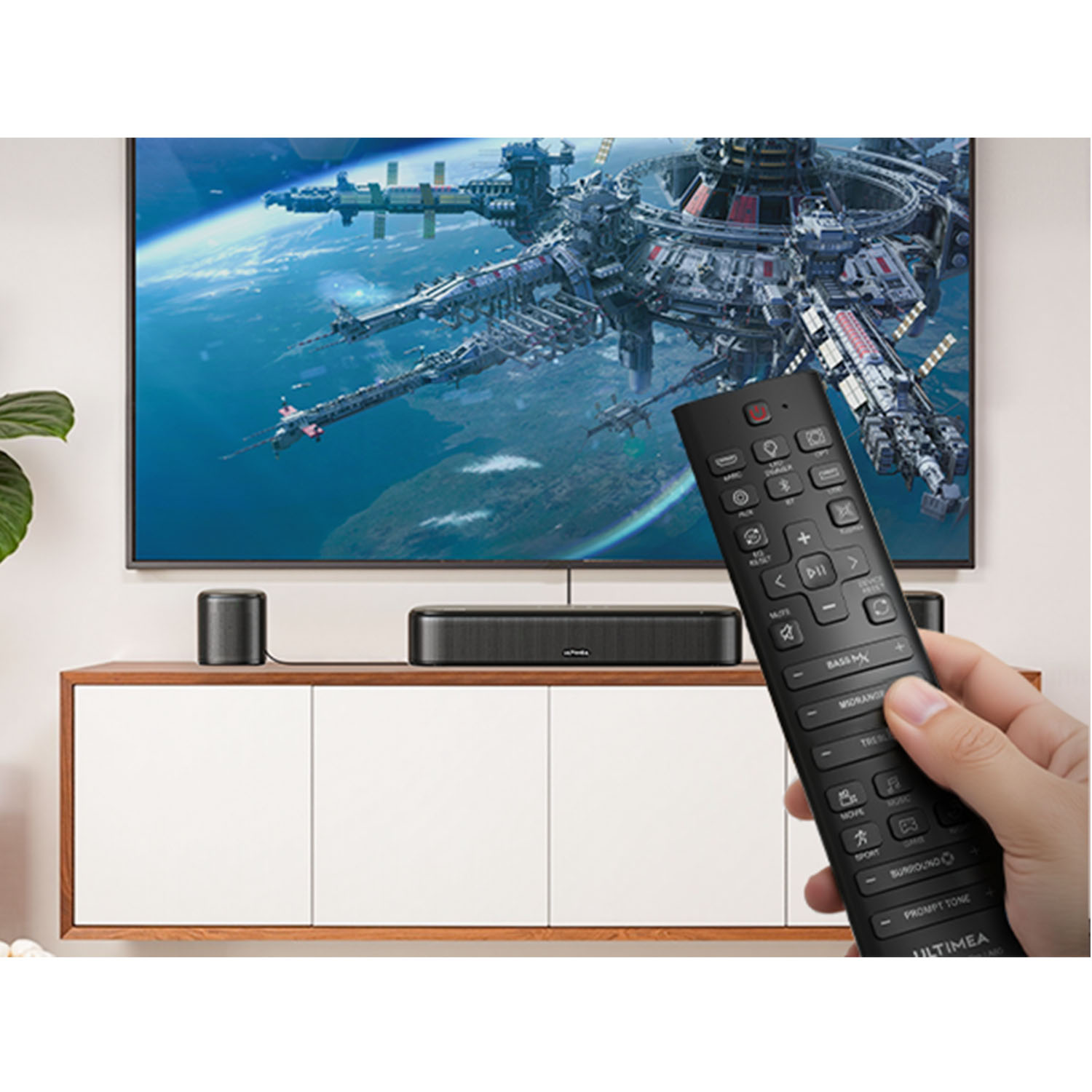 Ręka trzyma czarnego pilota przed telewizorem z kosmiczną stacją. Poniżej soundbar i głośnik.