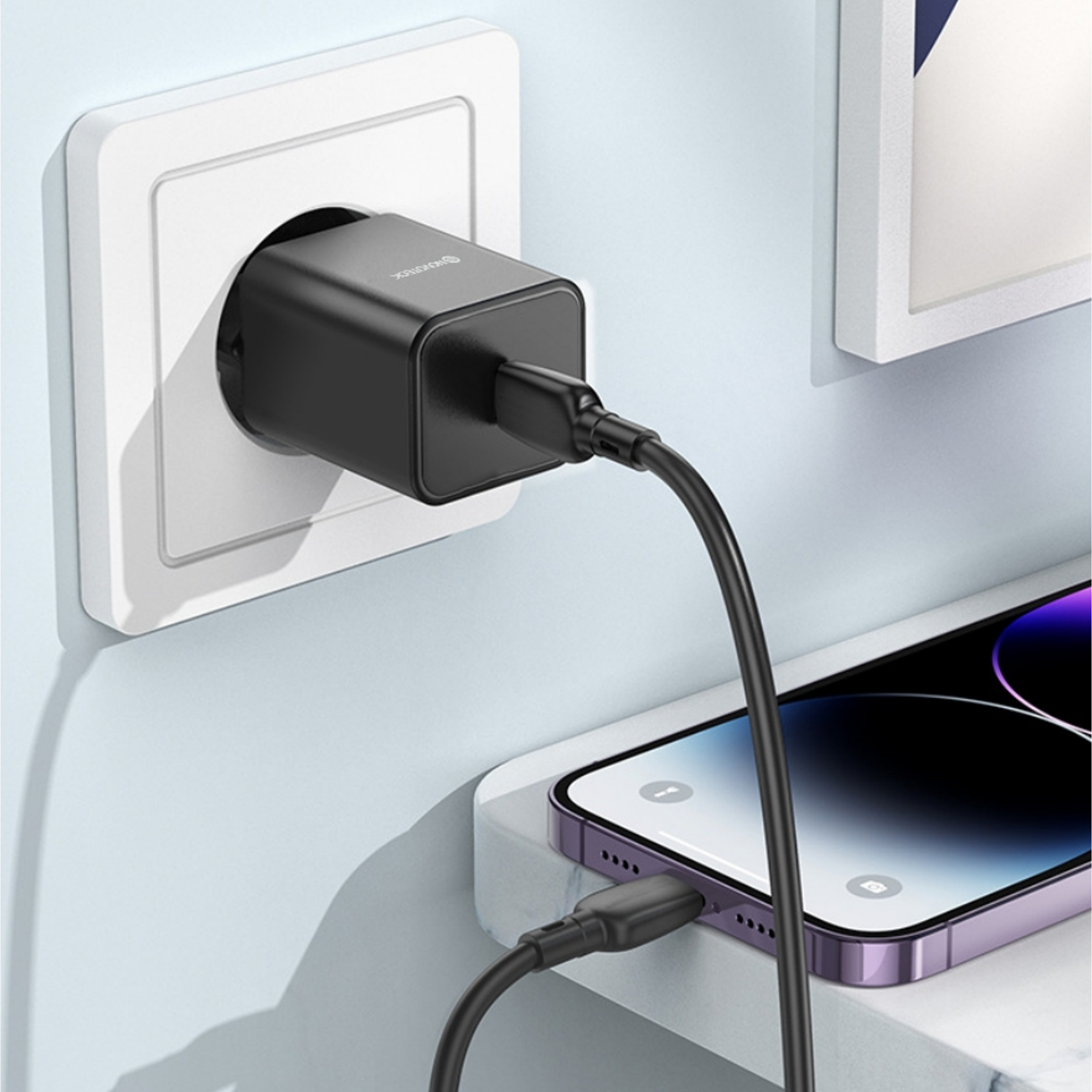 Cargador negro enchufado a una toma blanca, cargando un teléfono morado con un cable negro. Pared blanca al fondo.