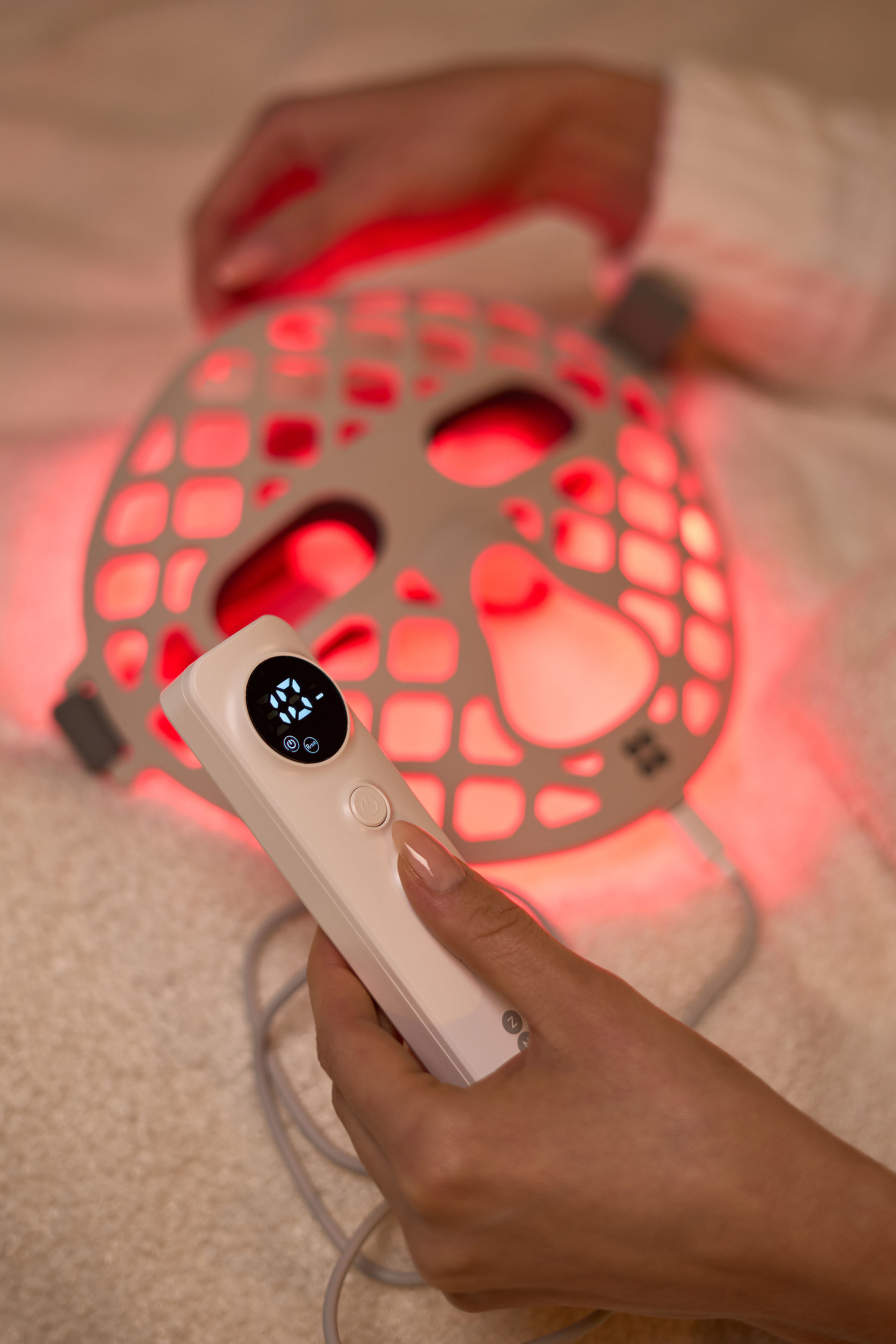 Osoba trzyma białe urządzenie z ekranem, w pobliżu czerwono oświetlonej maski na twarz. Tło jest białe.