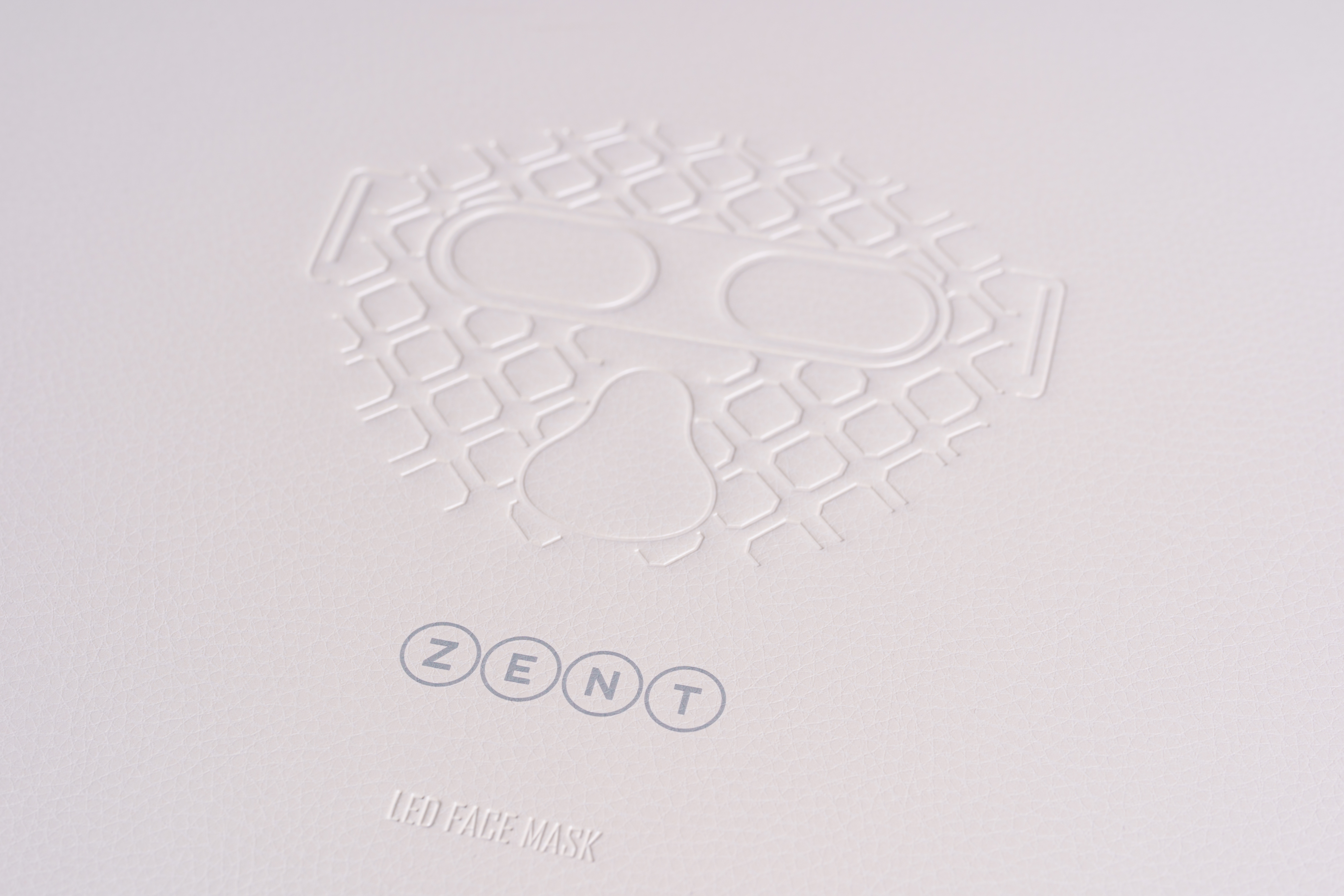 Biała powierzchnia z wytłoczonym wzorem maski na twarz, słowa 'ZENT' i 'LED FACE MASK' również wytłoczone.
