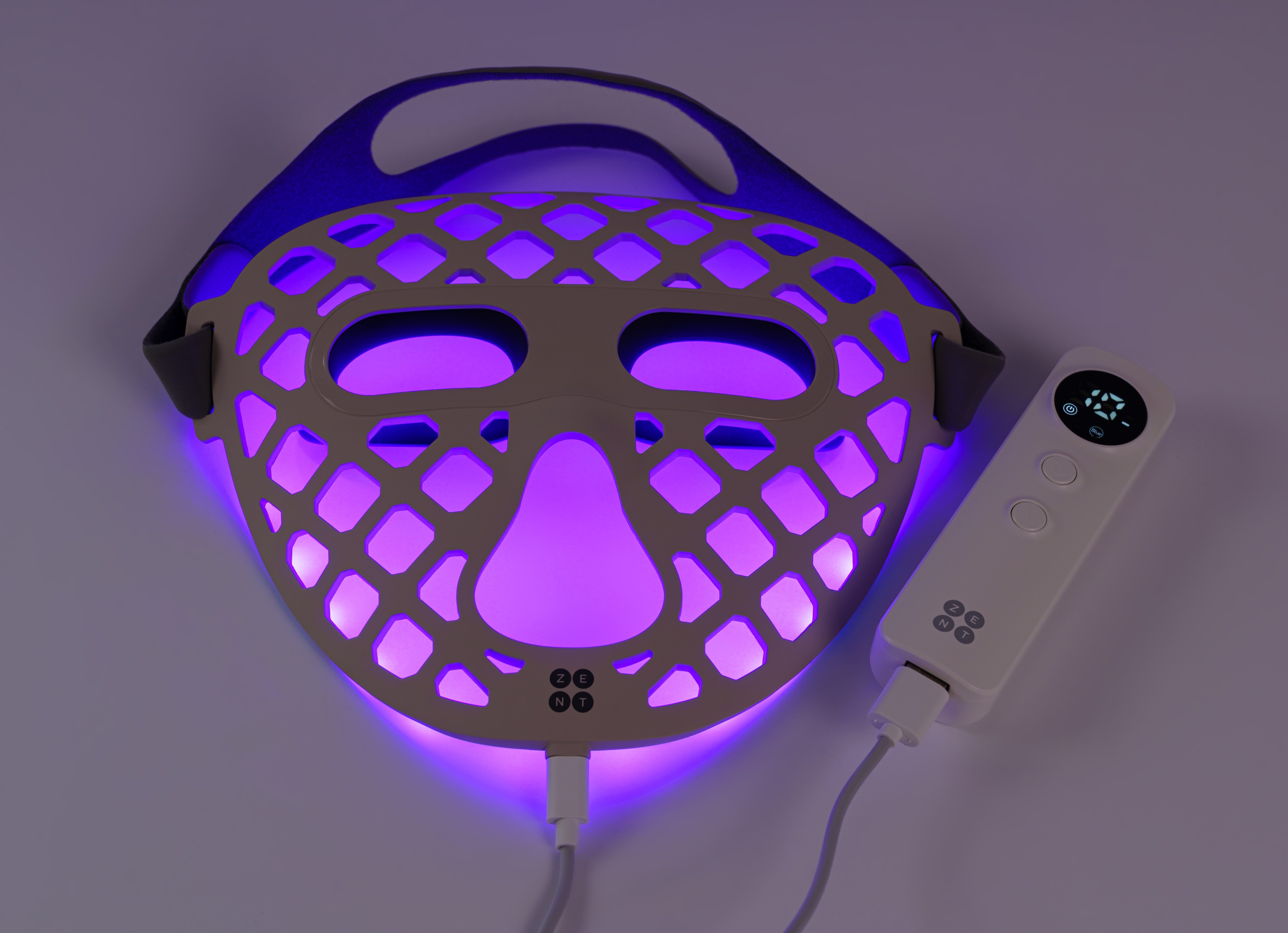 Maska na twarz z fioletowym światłem i białym kontrolerem, połączone kablem. Maska leży na białej powierzchni.