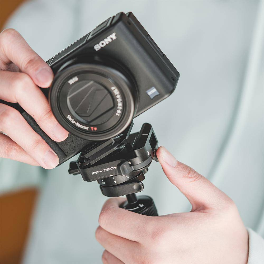 Eine Person hält eine schwarze Sony-Kamera, die an einem Stativkopf befestigt ist. Das Kameraobjektiv hat Zeiss-Markenzeichen. Hände sind sichtbar.
