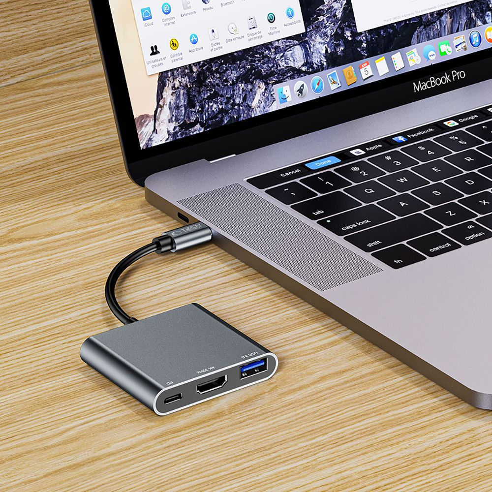 A gray USB hub is connected to a MacBook Pro on a wooden desk. The screen displays a webpage. The keyboard is in view.
