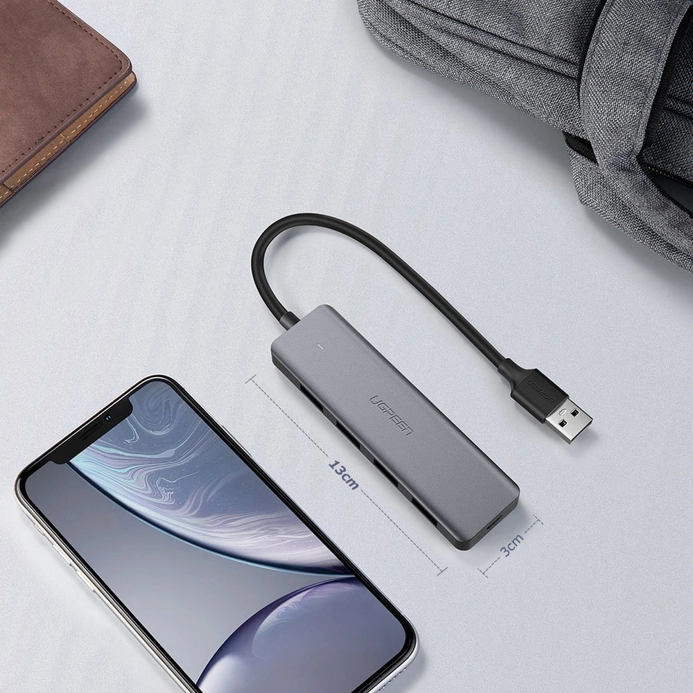 Un hub USB UGREEN grigio con cavo nero è collegato a un telefono. Un portafoglio marrone e una borsa grigia sono sullo sfondo.