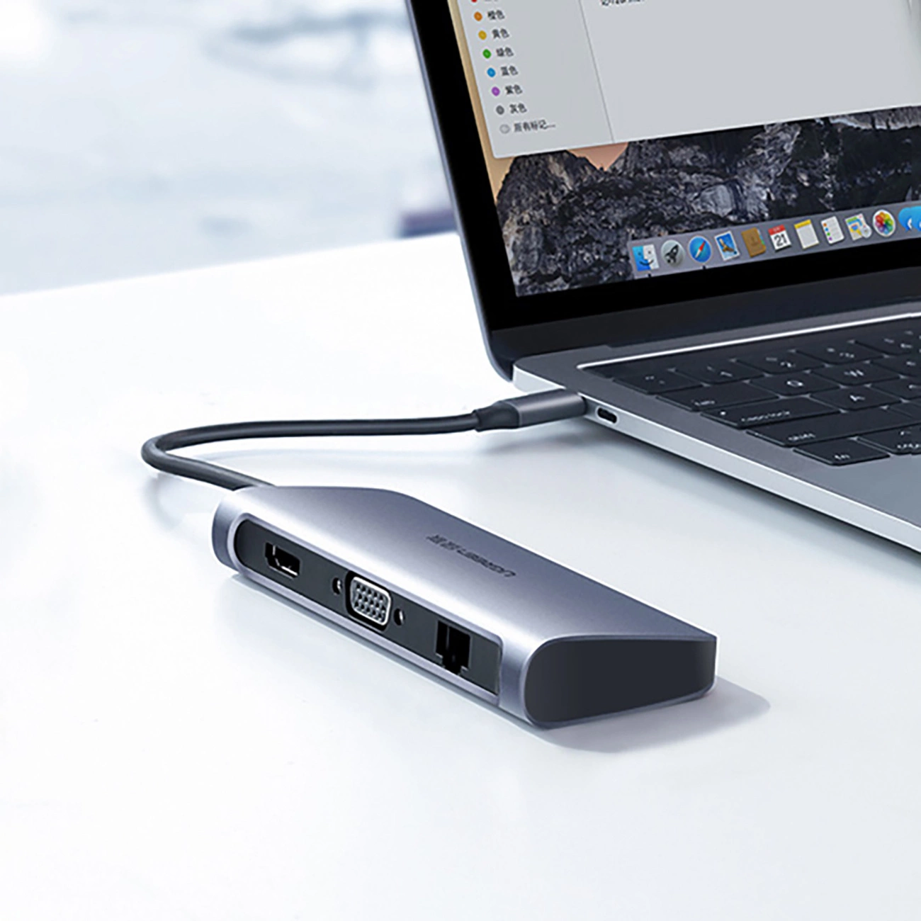 A gray USB hub is connected to a laptop. The laptop is open on a white surface. The hub has multiple ports.