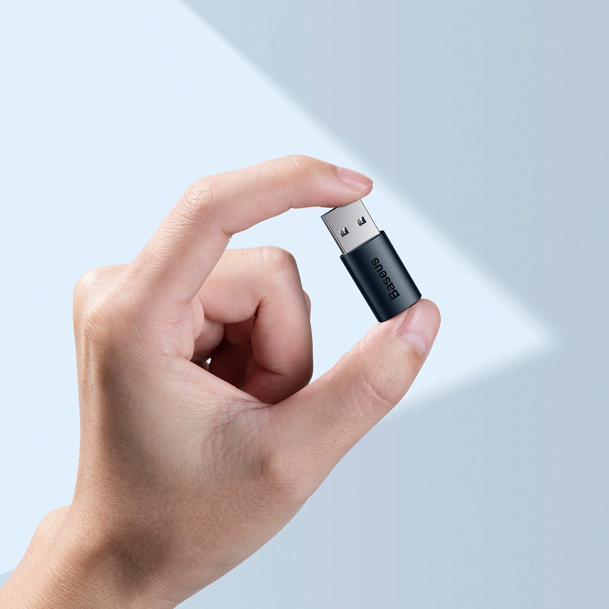 Dłoń trzyma czarny adapter USB ze srebrnym złączem USB. Widoczna jest nazwa marki 'Baseus'.