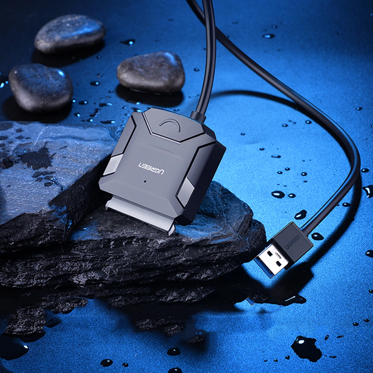 Szary adapter dysku twardego UGREEN z kablem USB na czarnej skale. Trzy kamienie w tle na niebieskiej powierzchni.