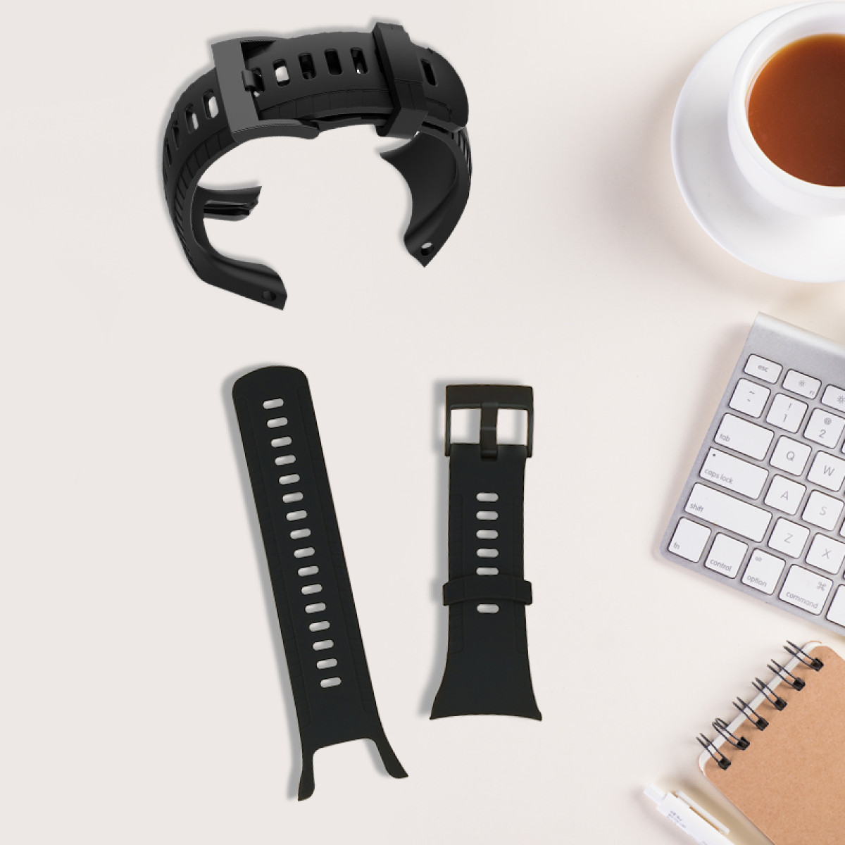 Cinturini e parti di orologio neri su una superficie bianca. Una tazza di caffè, una tastiera e un blocco note sono visibili.