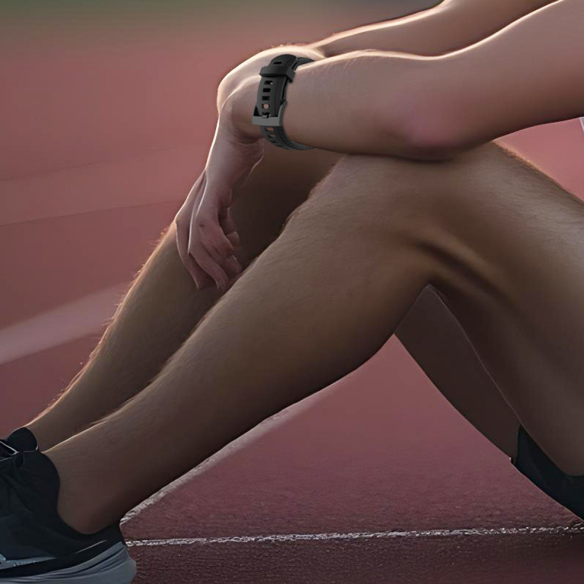Una persona seduta su una pista rossa, braccia attorno alle ginocchia. Un orologio nero al polso. Gambe e piedi visibili.