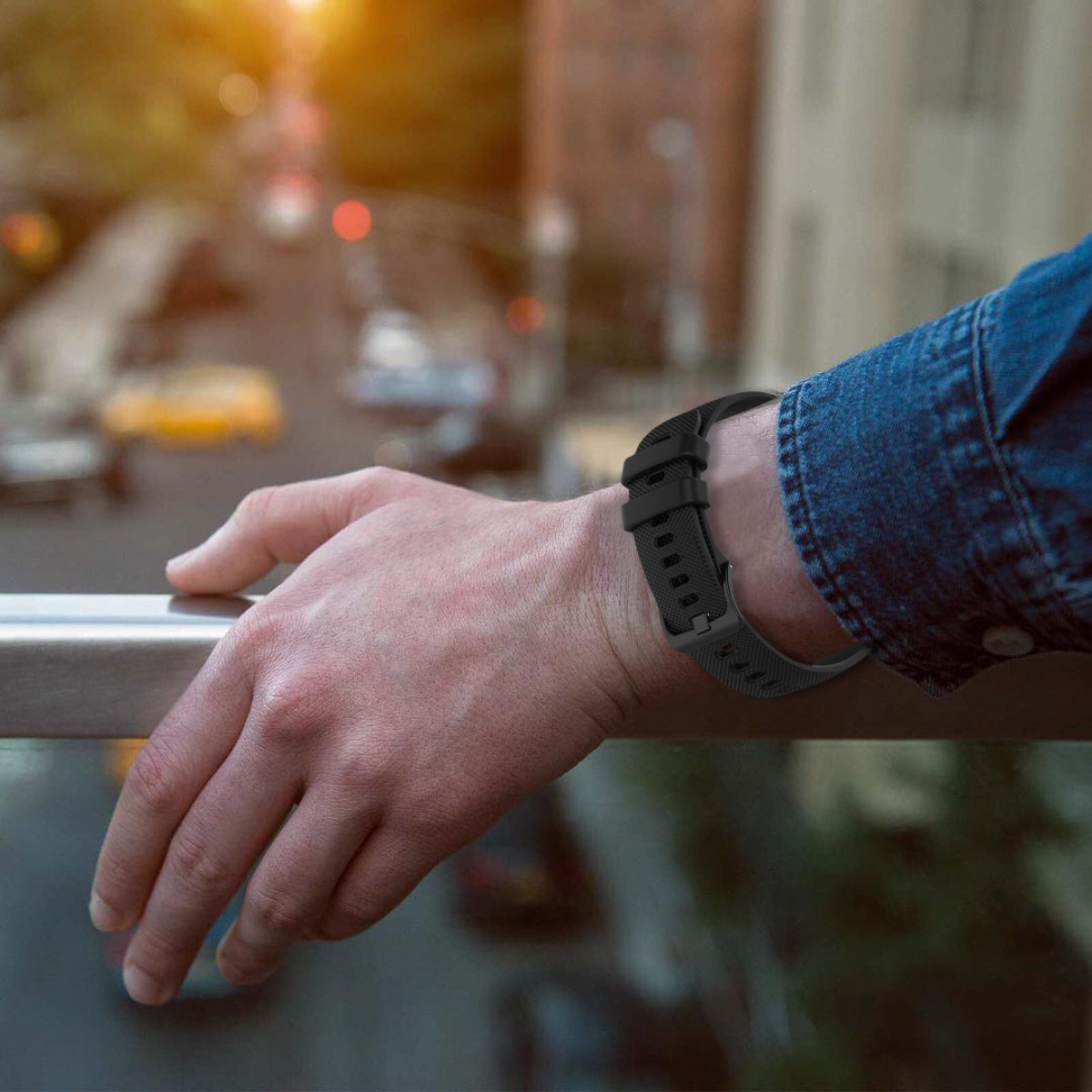 Ramię osoby oparte na balustradzie, noszące czarny zegarek. Samochody i budynki są rozmyte w tle, ze światłem słonecznym.