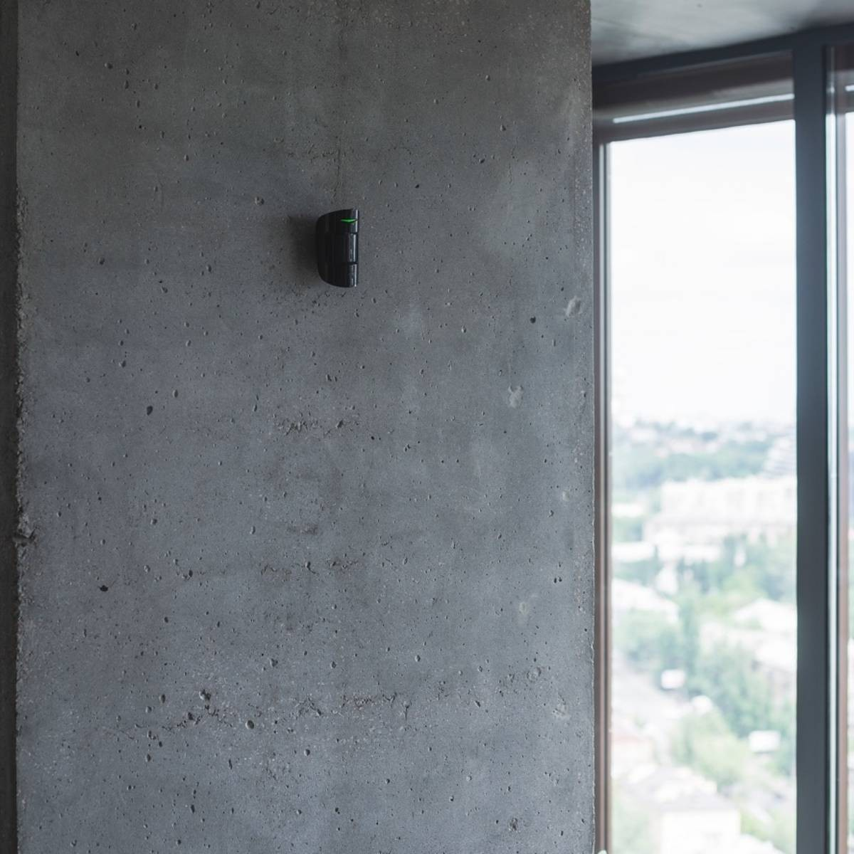 Un dispositivo oscuro con una luz verde está montado en una pared de hormigón gris. Una ventana enmarca un paisaje urbano.