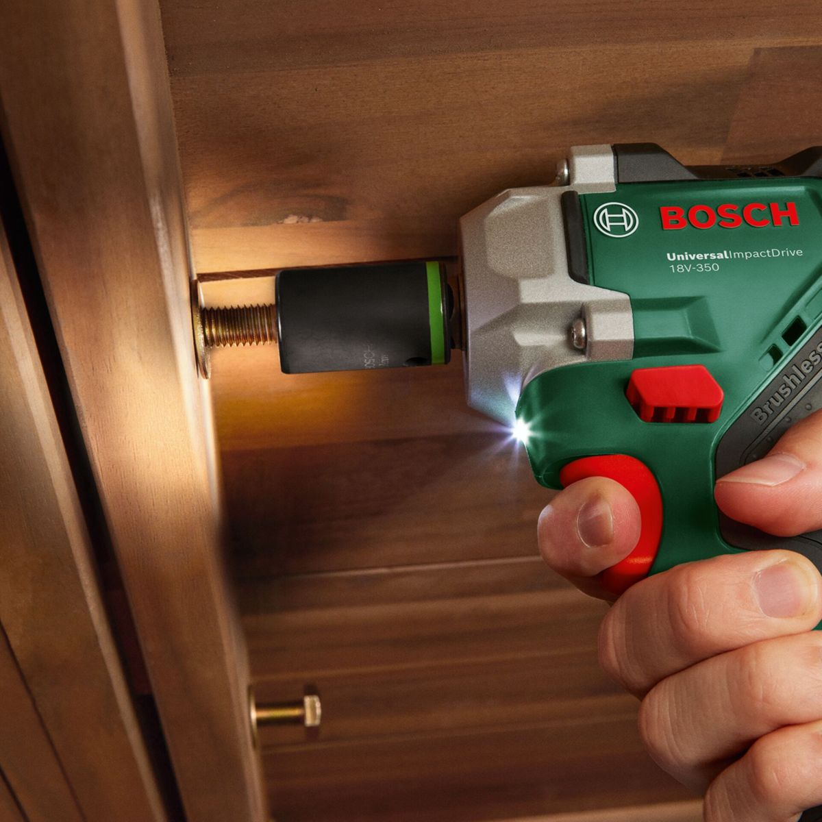 Ein grüner und grauer Bosch-Schlagschrauber schraubt eine Schraube in Holz. Eine Hand hält das Werkzeug. Die Arbeit wird vom Licht des Werkzeugs beleuchtet.