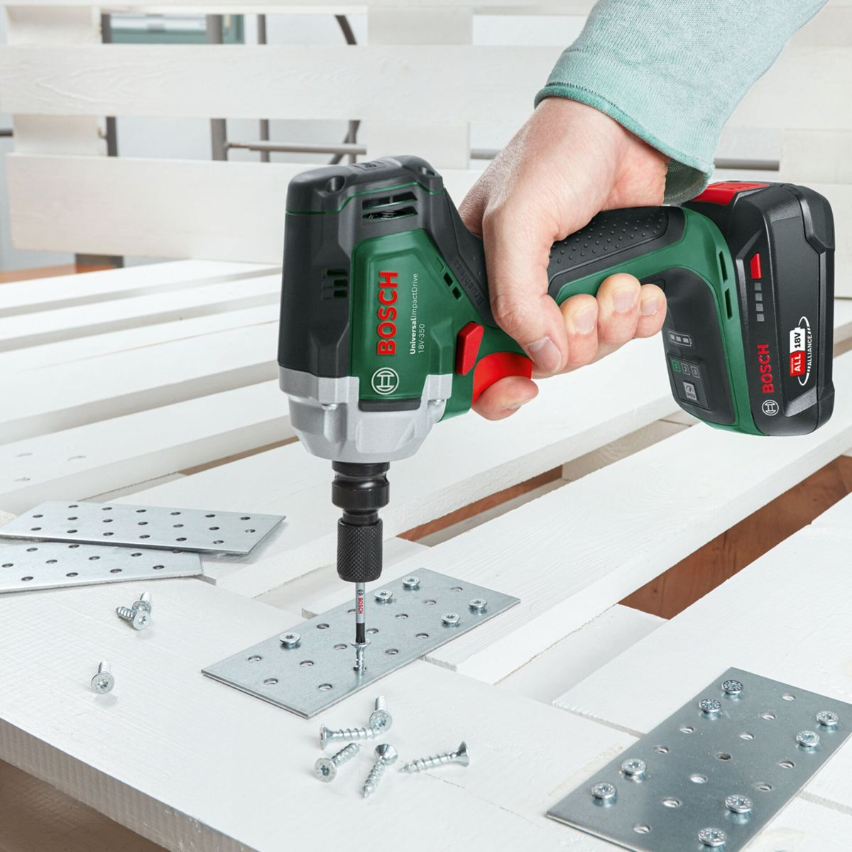Eine Hand hält einen Bosch-Schlagschrauber und schraubt eine Schraube in eine Metallplatte auf einer weißen Holzpalette. Andere Schrauben und Platten sind sichtbar.