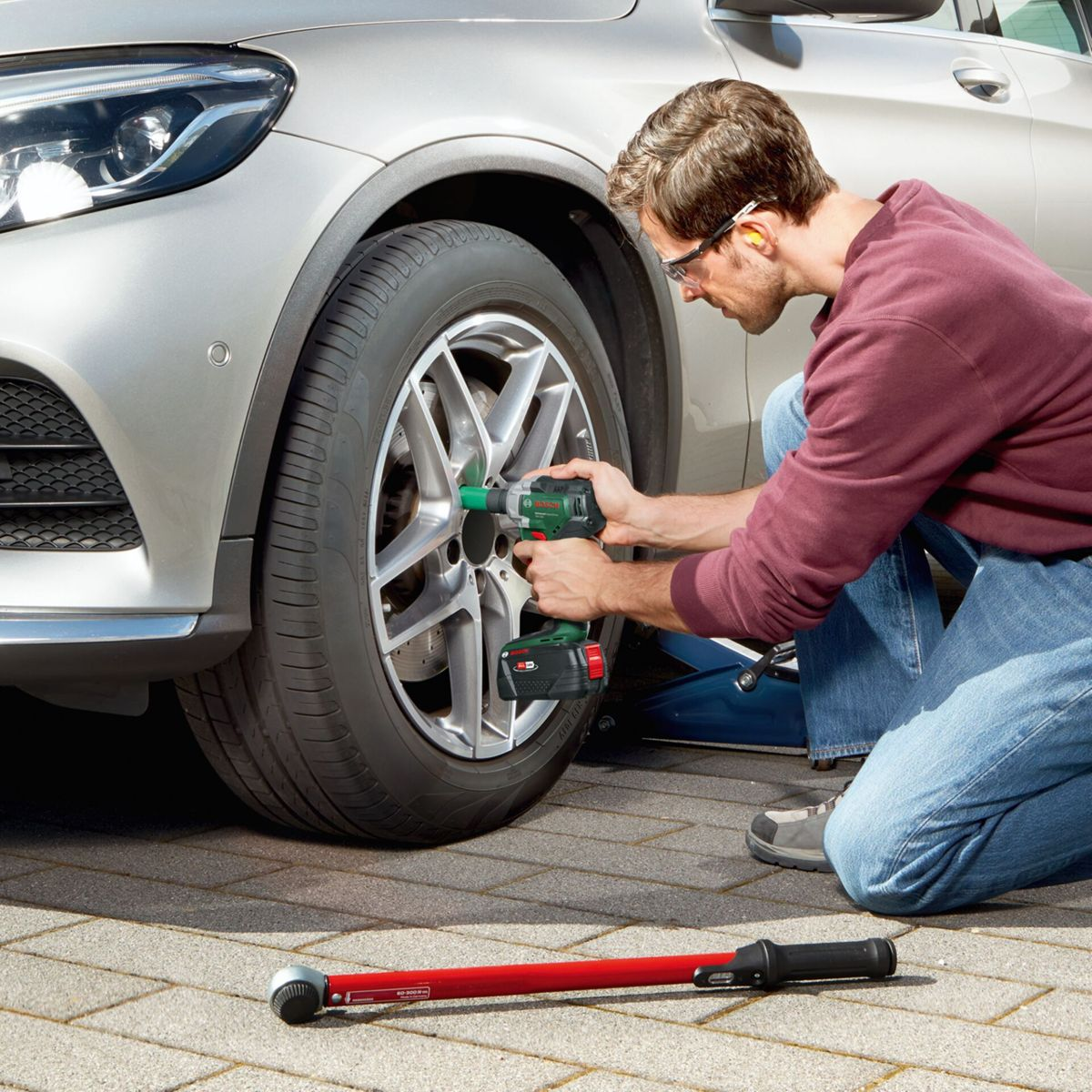Ein Mann in Jeans und einem weinroten Hemd benutzt ein Elektrowerkzeug an einem Autoreifen. Ein roter Drehmomentschlüssel liegt am Boden.