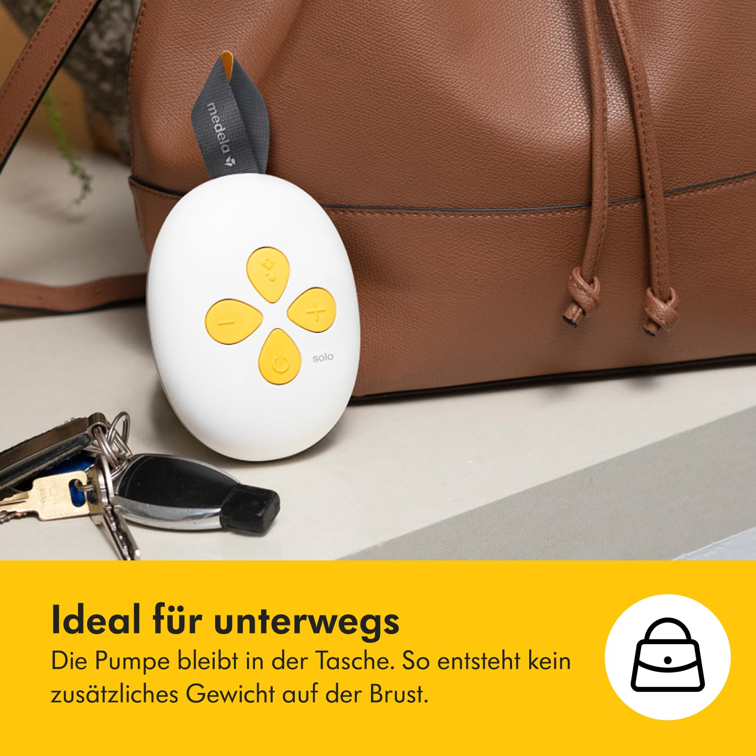 Eine weiße Brustpumpe mit gelben Knöpfen, eine braune Tasche und Schlüssel liegen auf einem Tisch. Die Pumpe hat einen Medela-Gurt.