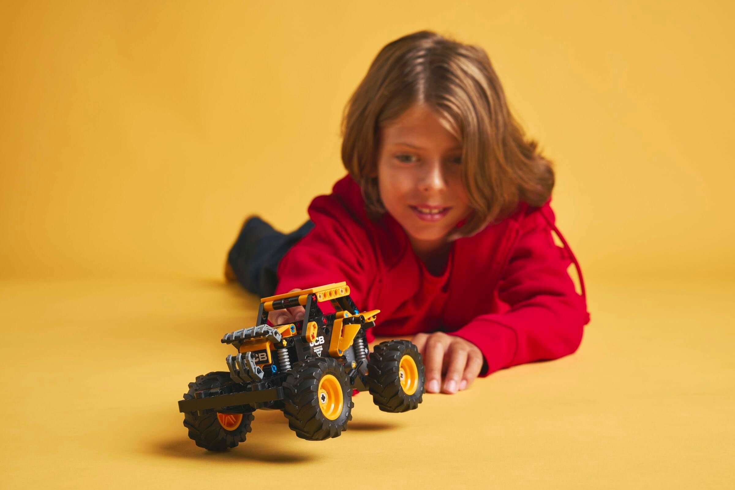 Ein Kind in einem roten Kapuzenpullover betrachtet einen Spielzeug-Monstertruck auf gelbem Hintergrund.