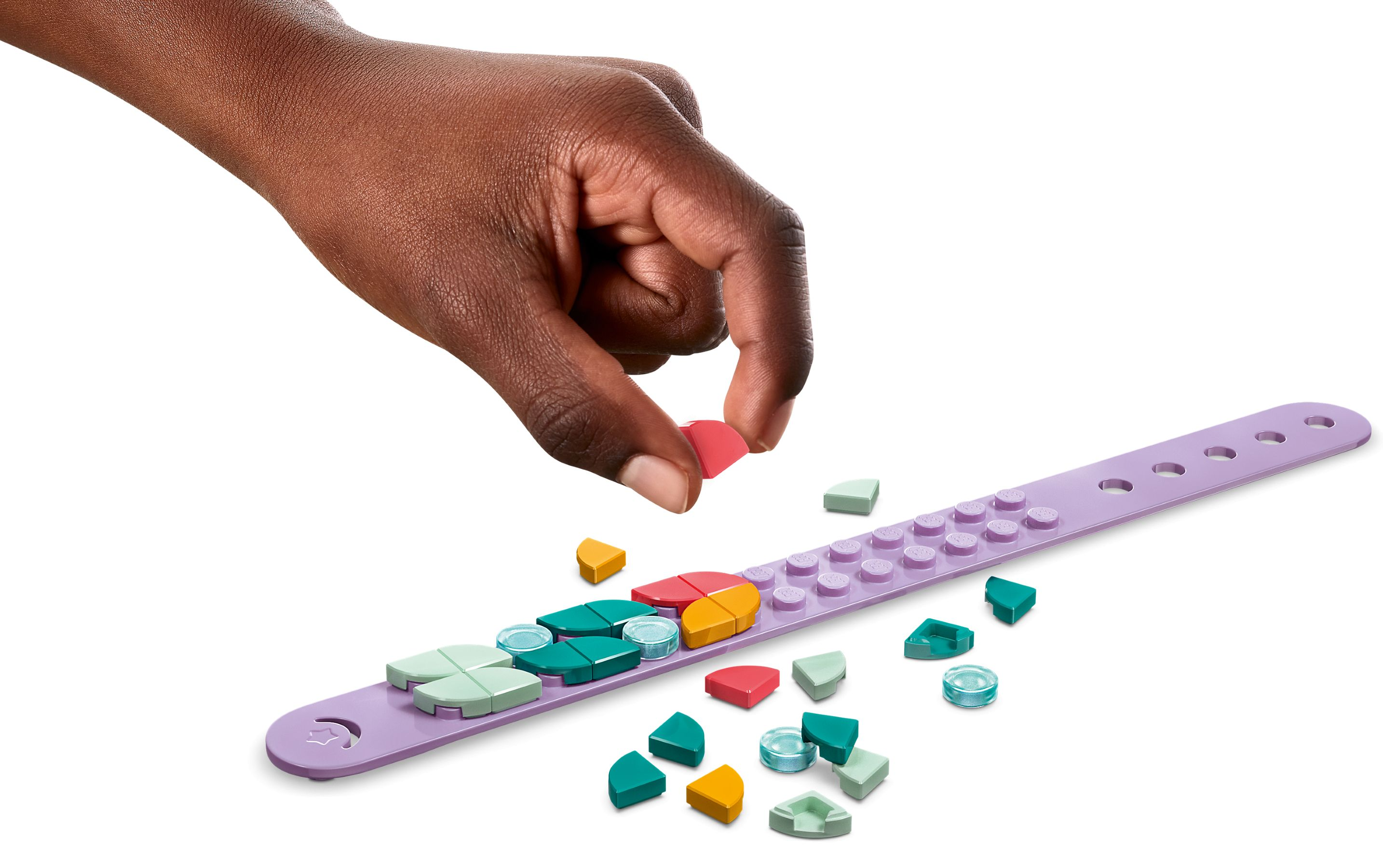 Eine Hand legt ein rotes LEGO-Teil auf ein lila Armband. Andere bunte Teile und ein weißer Hintergrund sind vorhanden.