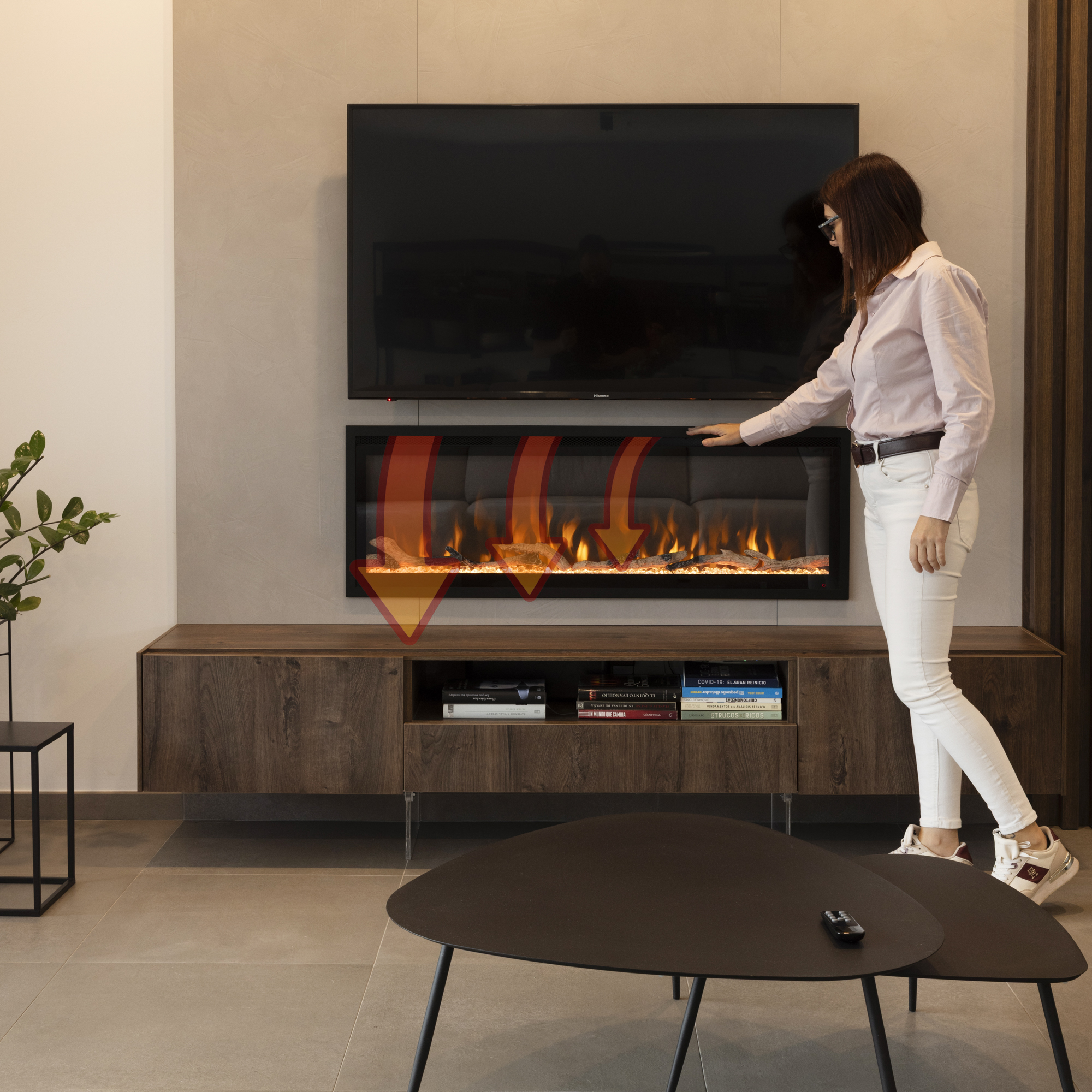 Una mujer toca una chimenea debajo de un televisor. Flechas rojas indican calor. Libros y una mesa en primer plano.