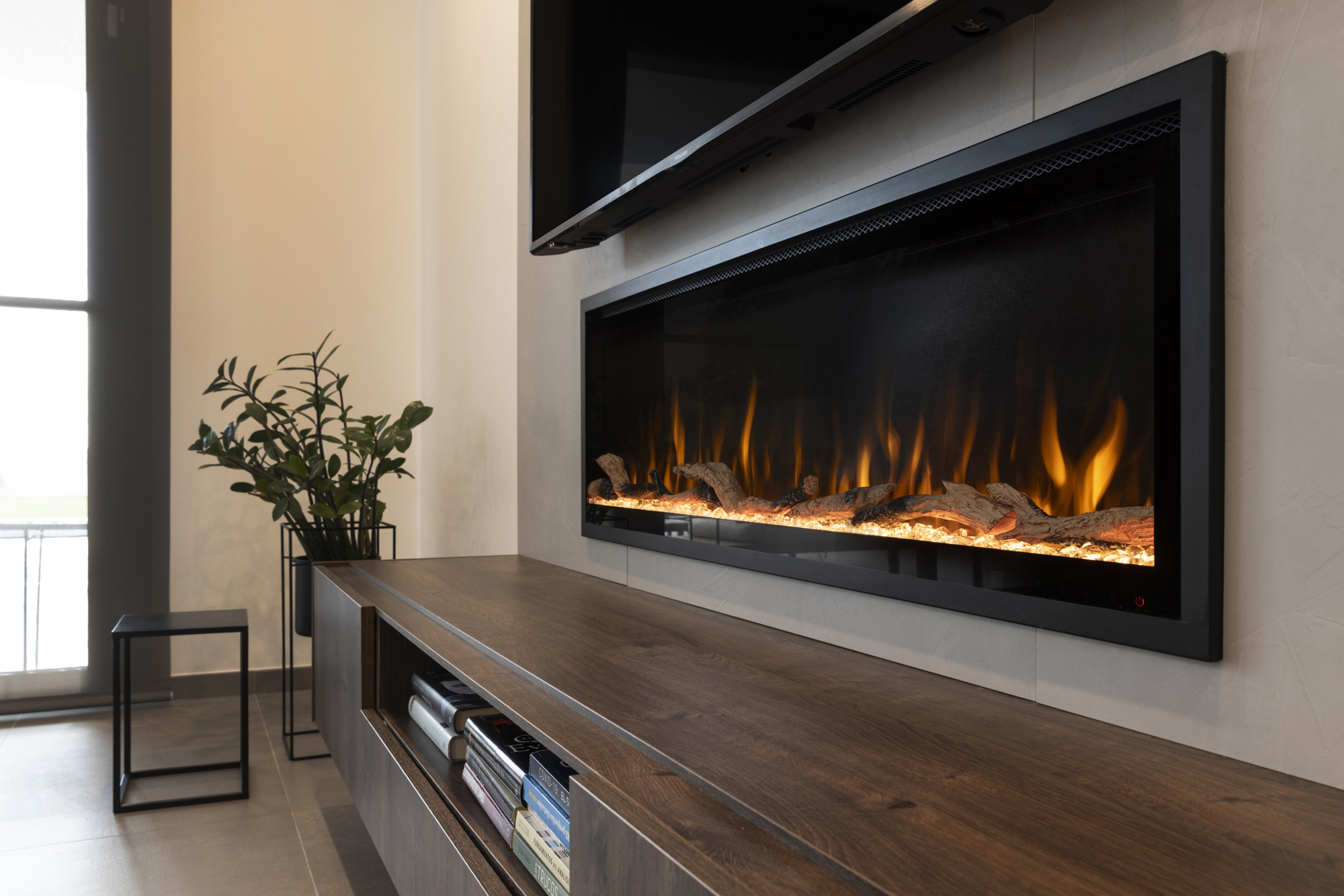 Una sala de estar moderna con chimenea, televisor y mueble de madera. Las llamas parpadean en la chimenea. Hay una planta y una mesita.