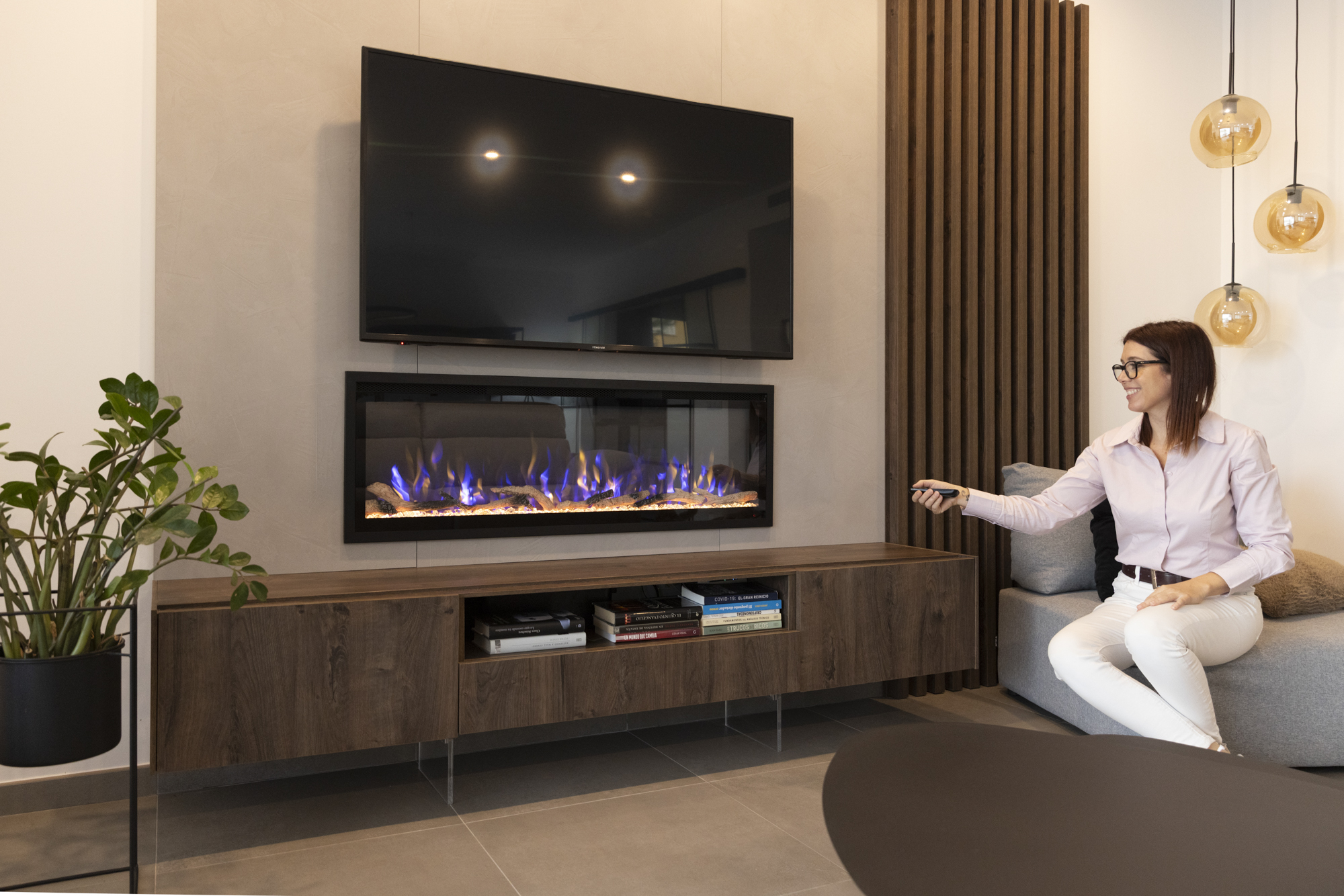 Una mujer con gafas y pantalones blancos usa un mando a distancia. Detrás hay un televisor sobre una chimenea, libros y una planta.