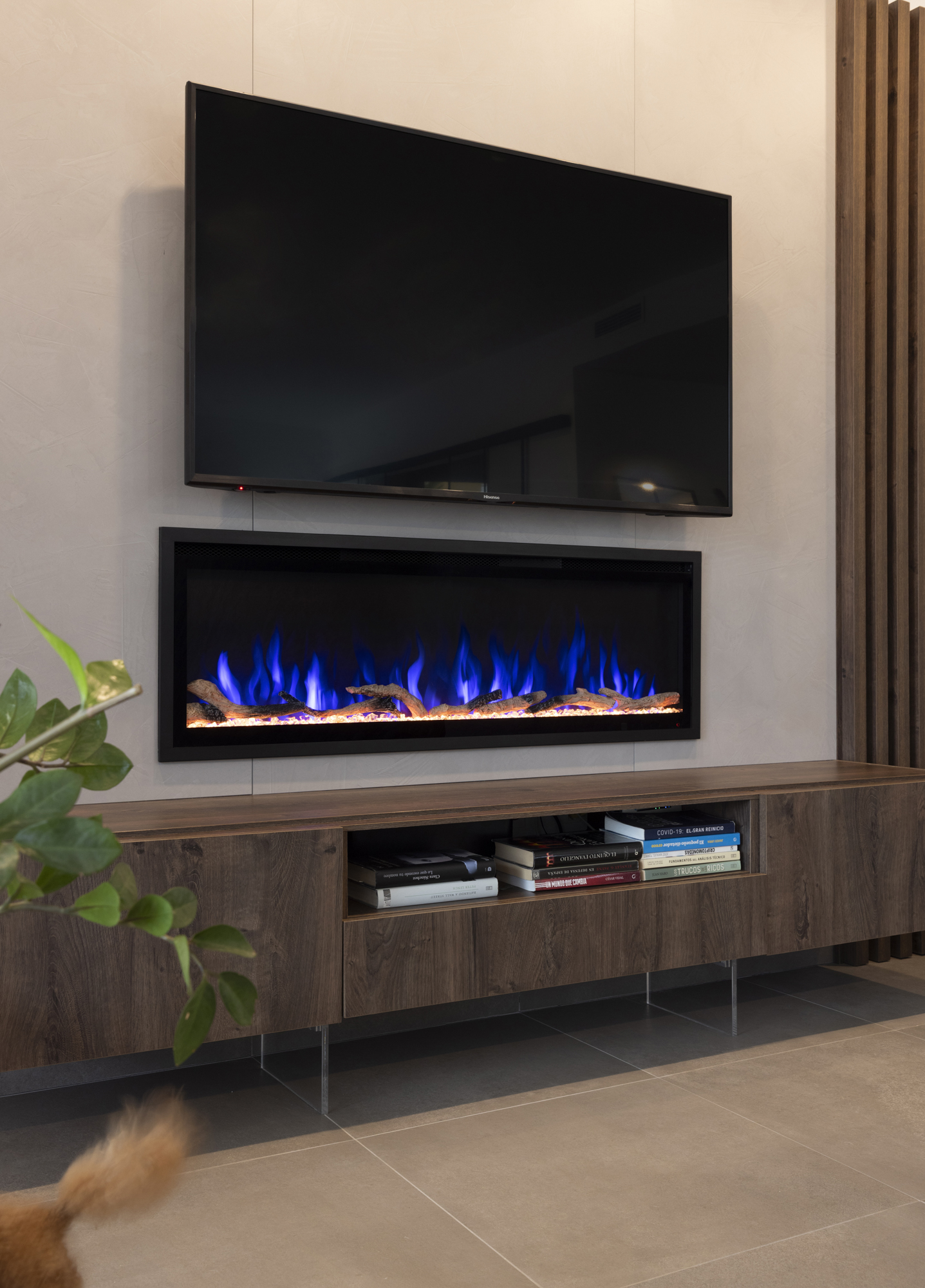 Un televisor se encuentra sobre una chimenea con llamas azules. Un armario de madera contiene libros. Una planta está en primer plano.