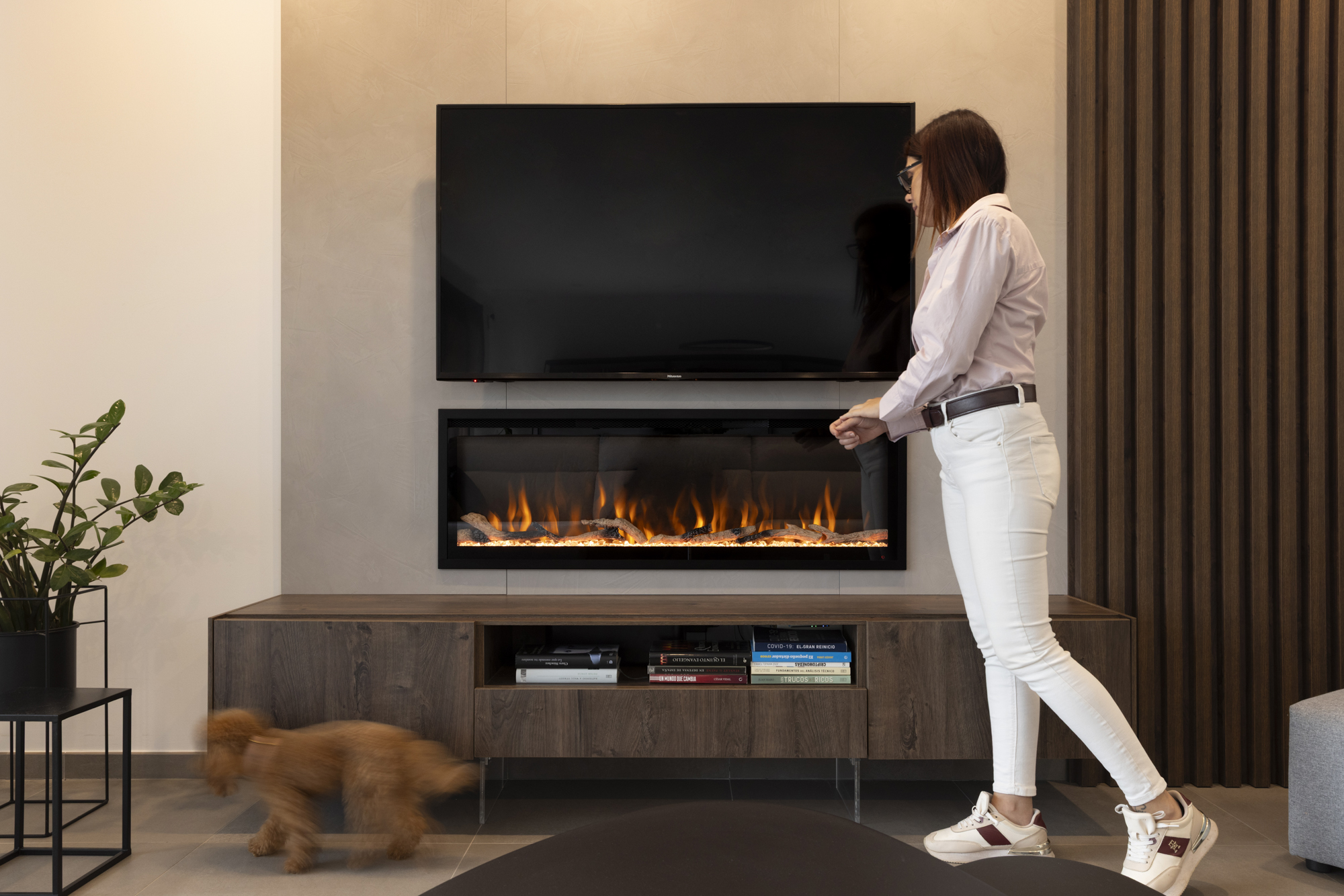 Una mujer está de pie cerca de una chimenea con un televisor encima. Un perro borroso corre junto a un armario con libros.