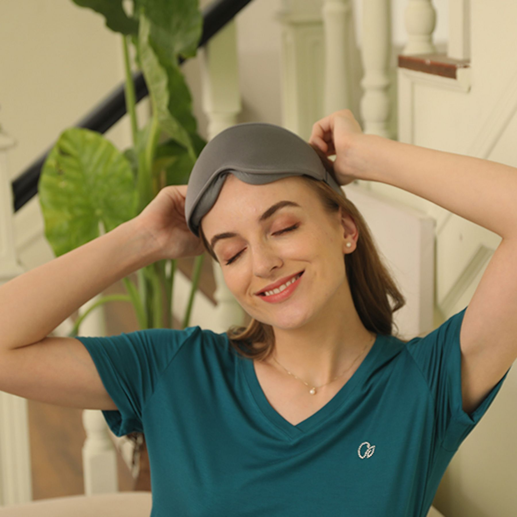 Frau mit geschlossenen Augen setzt eine graue Schlafmaske auf. Sie trägt ein türkisfarbenes Hemd und lächelt.