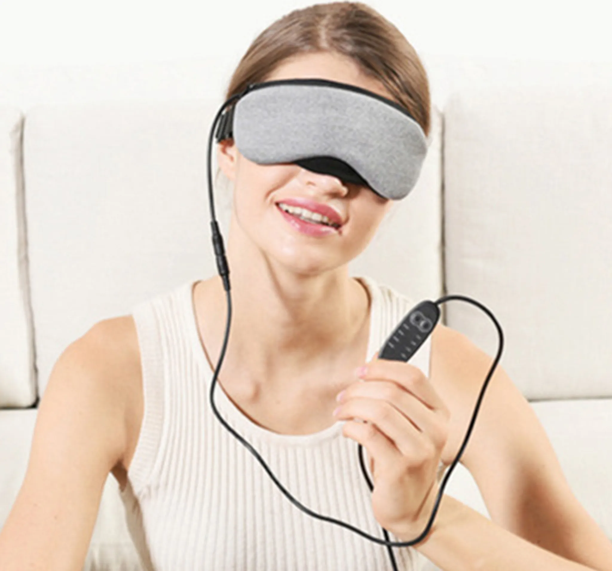Eine Frau mit grauer Augenmaske hält einen Controller. Sie lächelt. Der Hintergrund ist ein weißes Sofa.