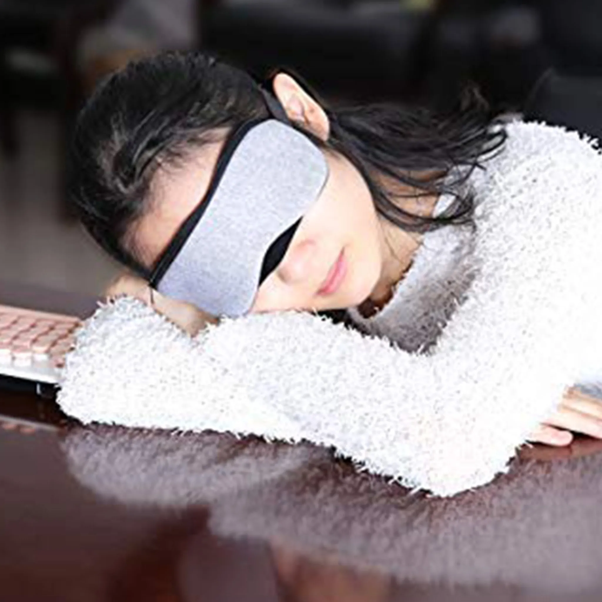 Eine Frau mit grauer Schlafmaske ruht ihren Kopf auf einem Tisch. Sie trägt einen weißen Pullover. Eine Tastatur ist sichtbar.