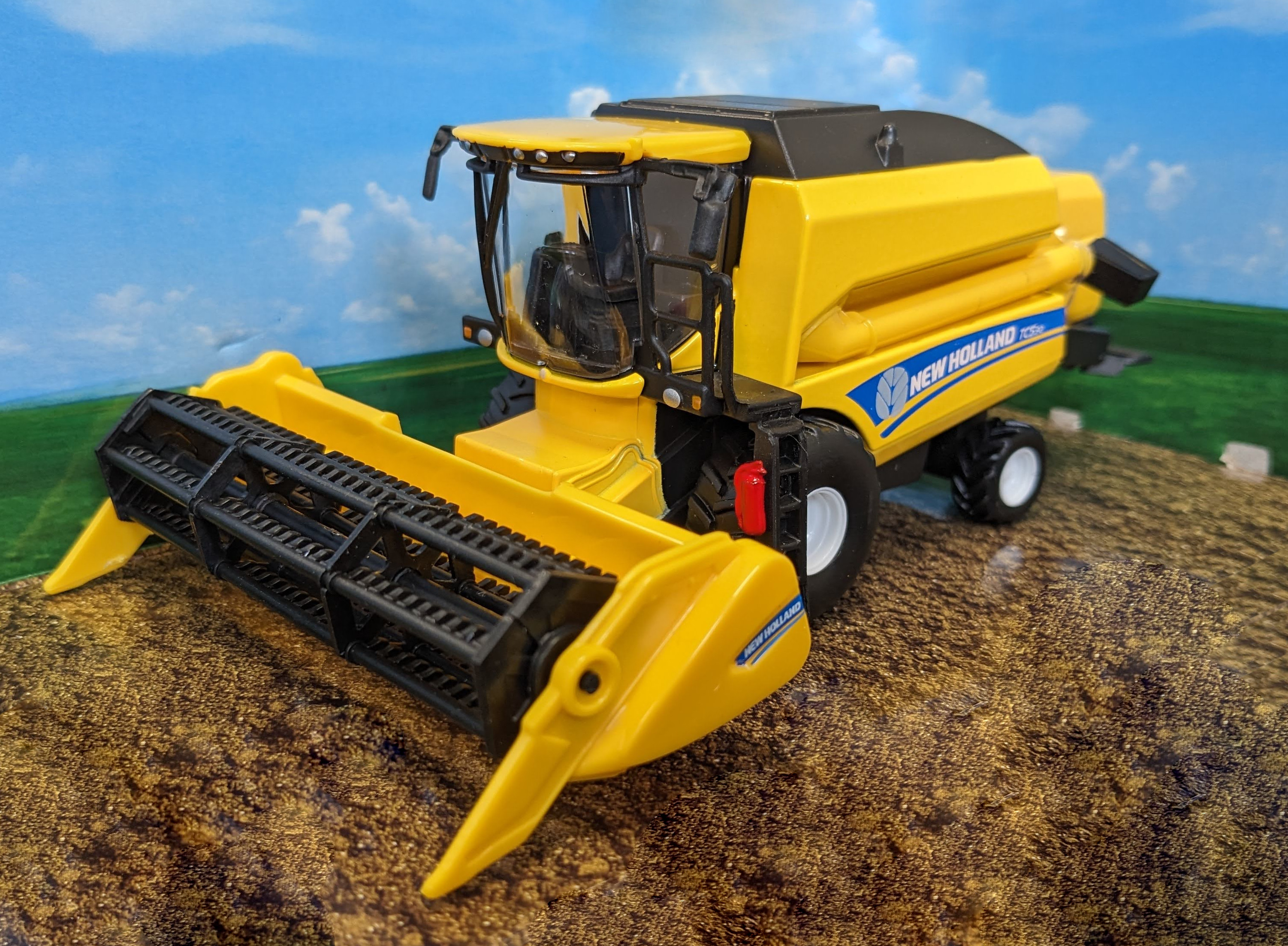 Gelbes New Holland Mähdrescher-Spielzeug auf brauner Oberfläche, vor blauem Himmel und grünem Feldhintergrund.