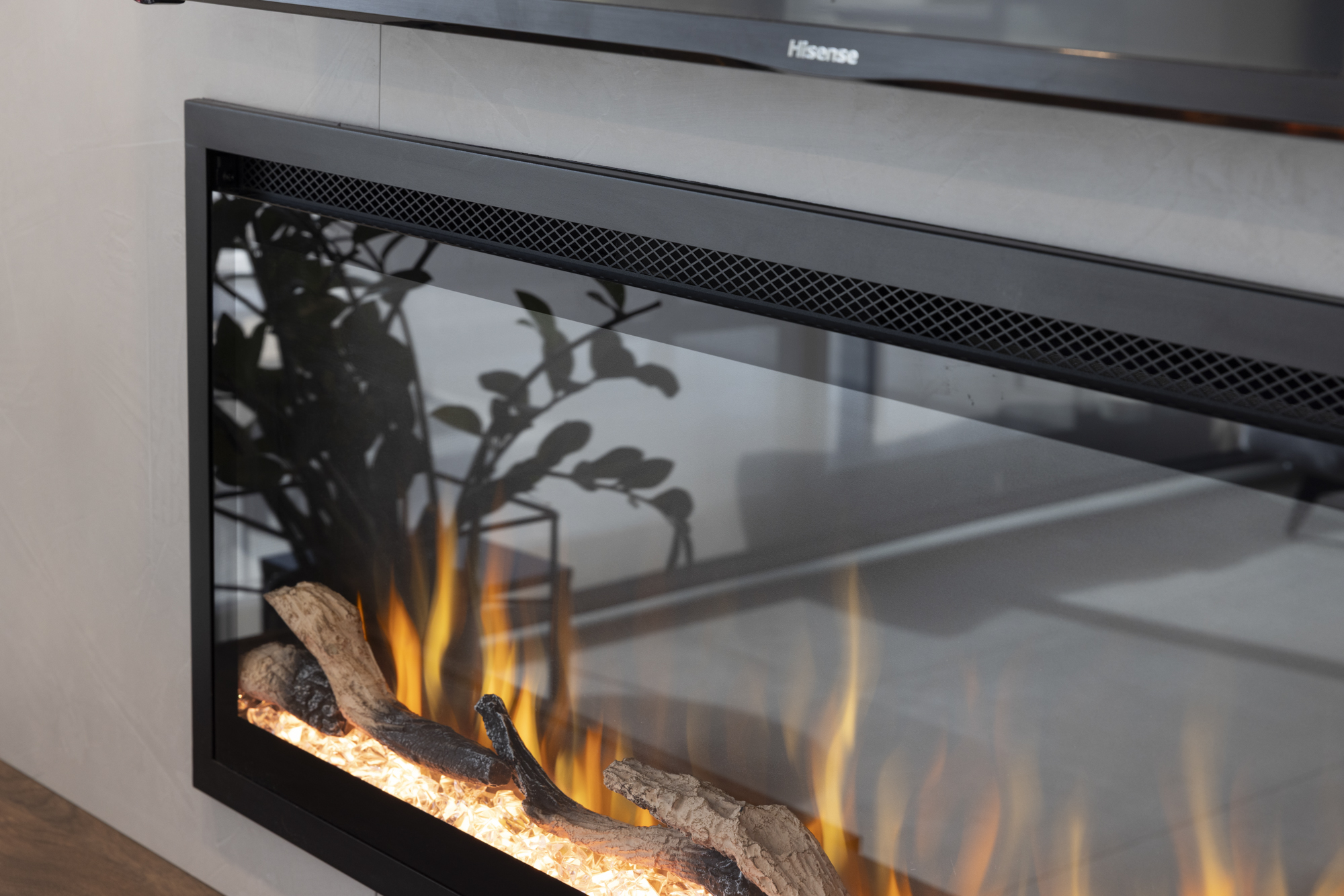 Chimenea eléctrica con llamas y troncos realistas, detrás de cristal. Un marco negro, un televisor Hisense y una silueta de planta son visibles.