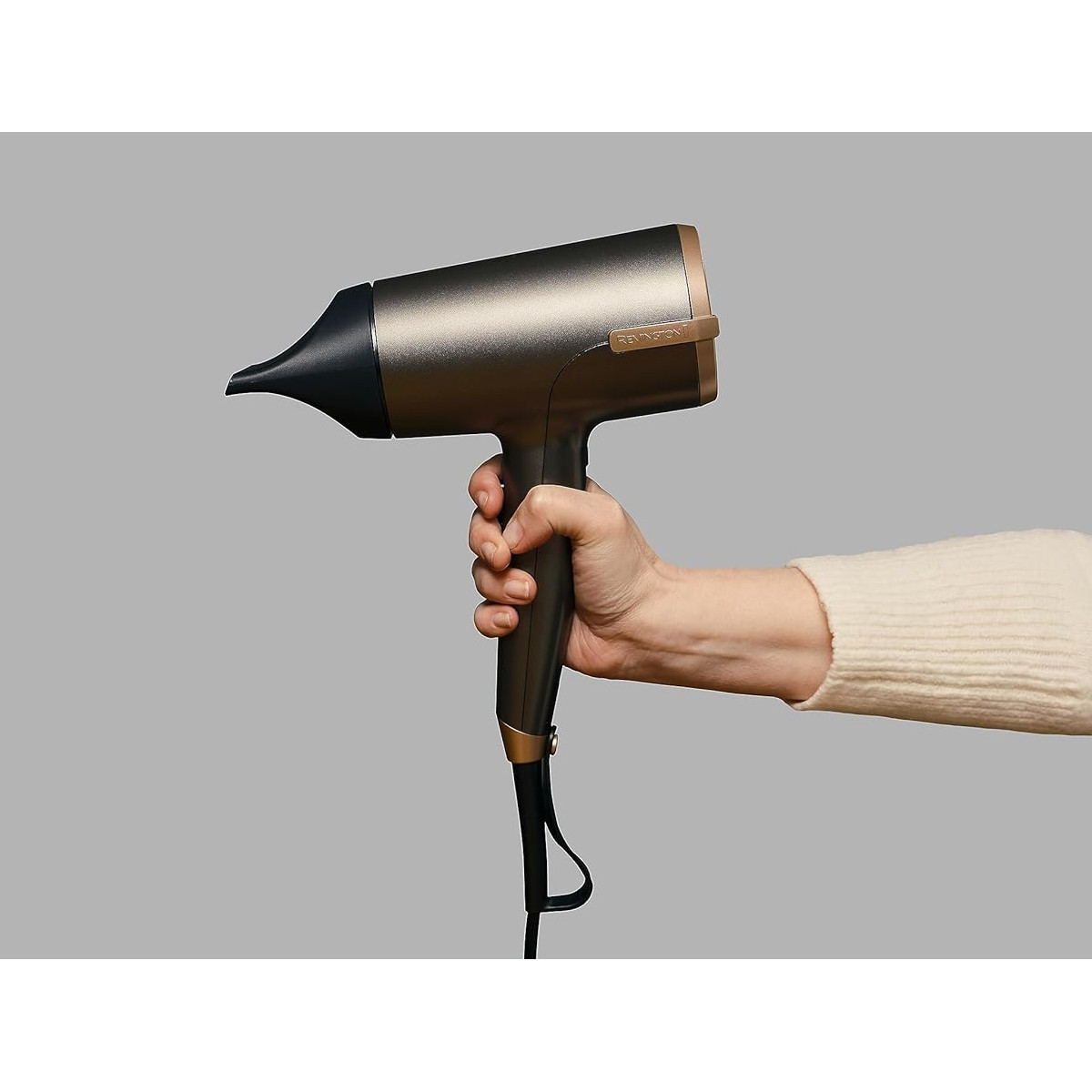 Una mano tiene un asciugacapelli grigio e oro. Lo sfondo è grigio. La persona indossa un maglione color crema.