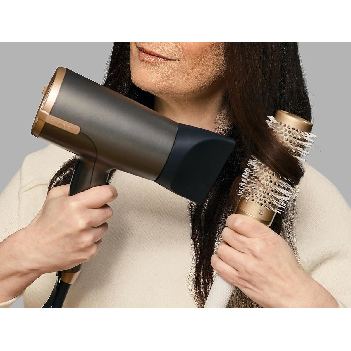 Una donna usa un asciugacapelli e una spazzola rotonda sui suoi capelli scuri. L'asciugacapelli è nero e oro.