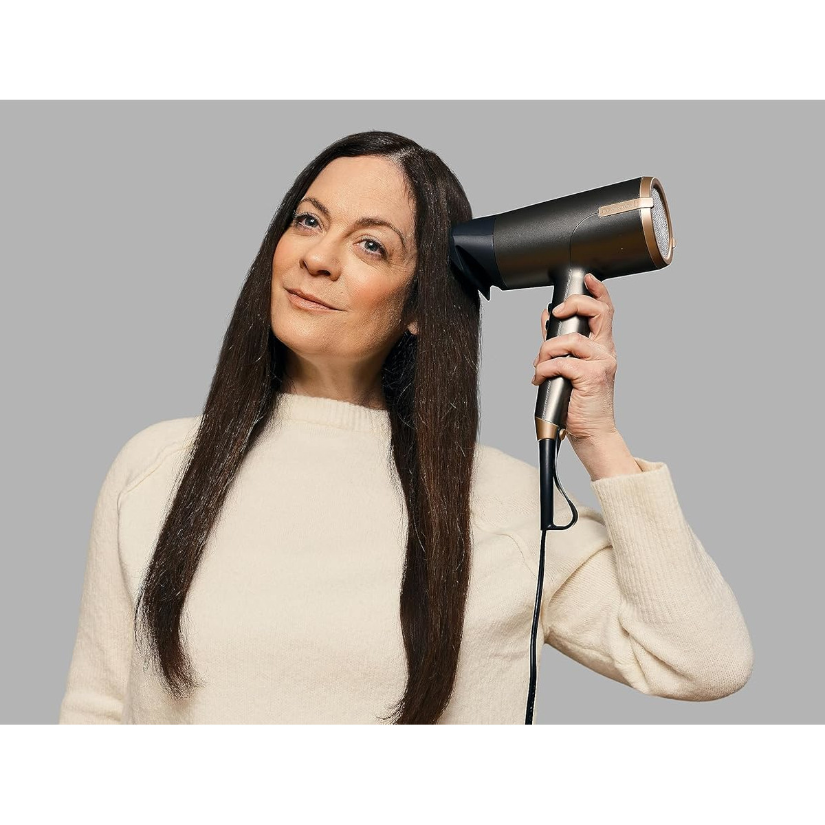 Una donna con lunghi capelli scuri usa un asciugacapelli. Indossa un maglione color crema su sfondo grigio.