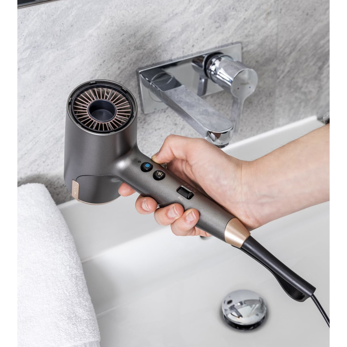 Eine Hand hält einen grauen Haartrockner über einem weißen Waschbecken. Ein Wasserhahn und ein weißes Handtuch sind ebenfalls zu sehen.