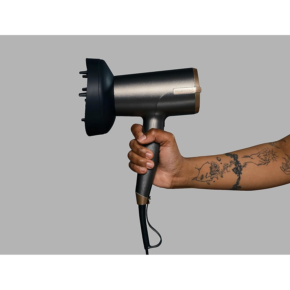 Una mano tiene un asciugacapelli grigio scuro con un diffusore nero. Il braccio ha tatuaggi su sfondo grigio.