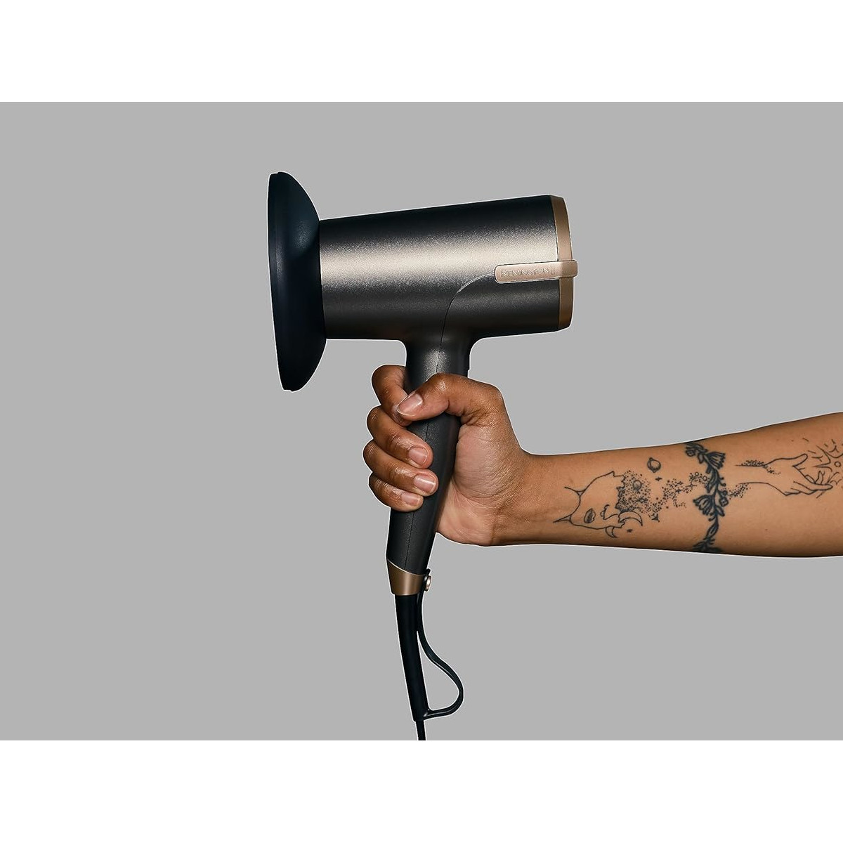 Una mano tiene un asciugacapelli scuro con dettagli dorati. Il braccio ha tatuaggi. Lo sfondo è grigio.