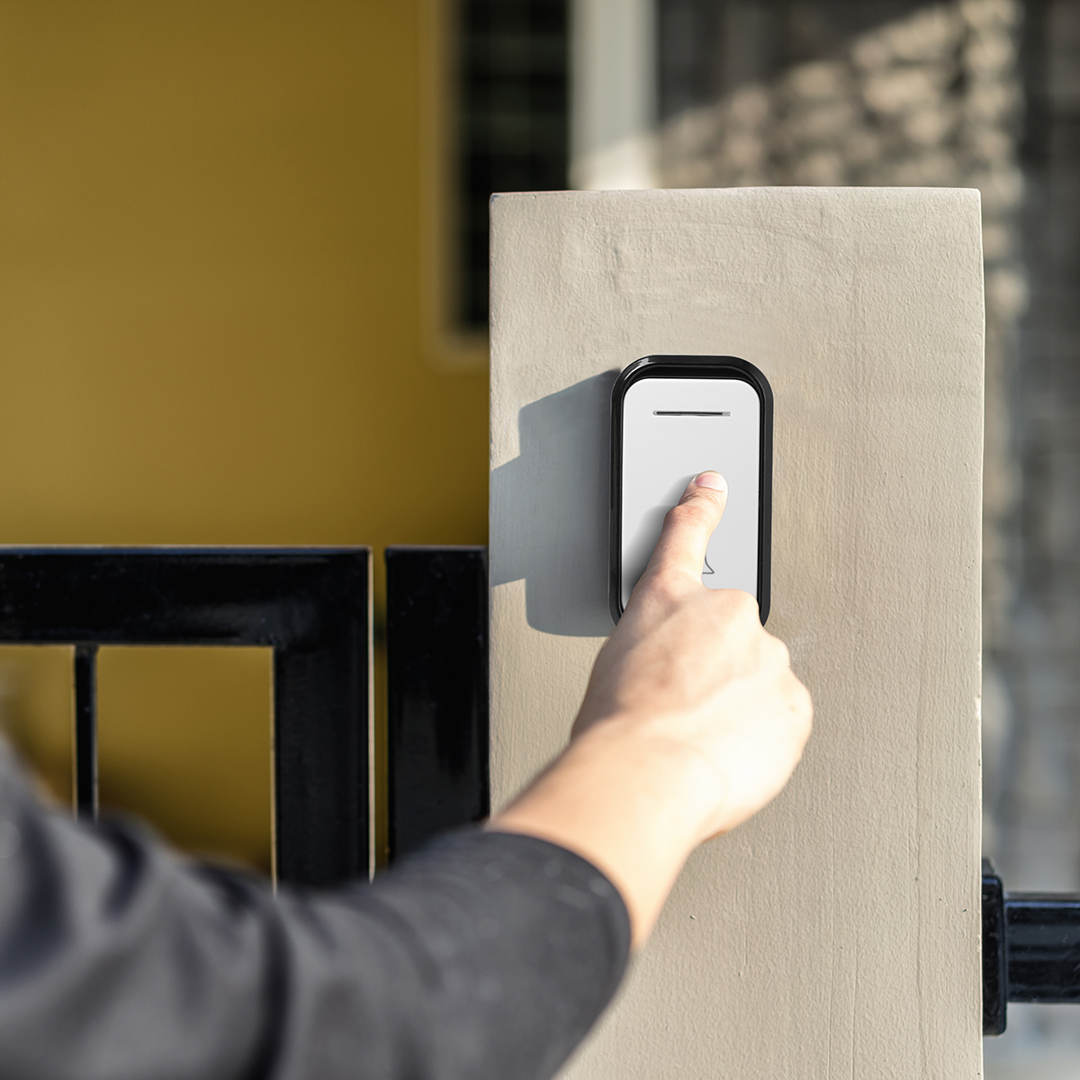 Ręka naciska dzwonek do drzwi na szarym filarze. Za nim żółta ściana i czarna brama. Światło słoneczne rzuca cienie.