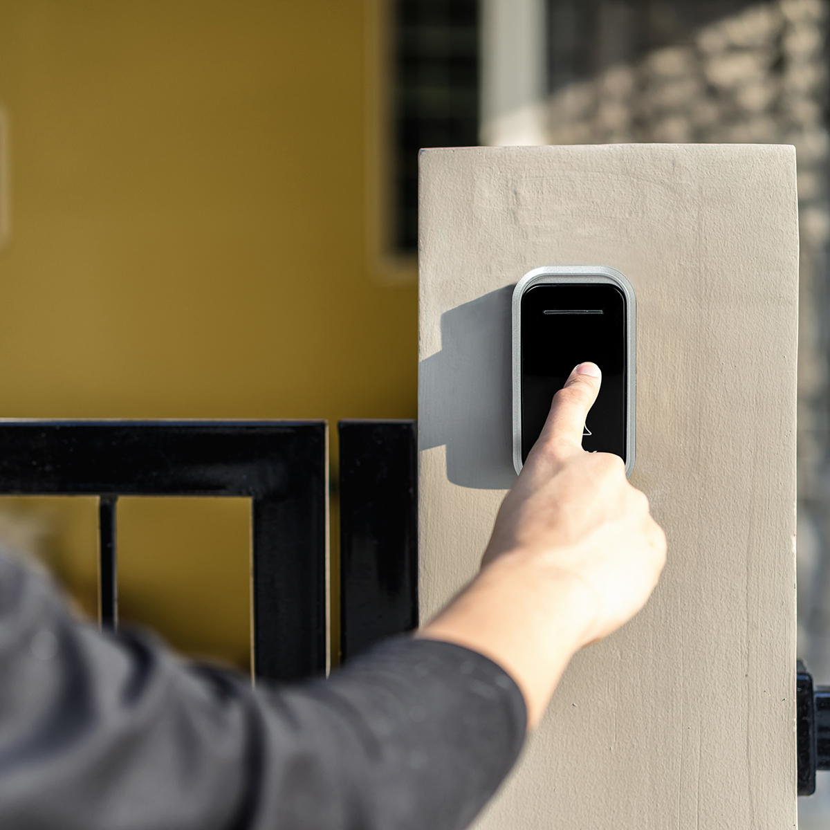 Dłoń naciska czarny przycisk dzwonka na beżowym filarze. Czarne ogrodzenie i żółta ściana są w tle.