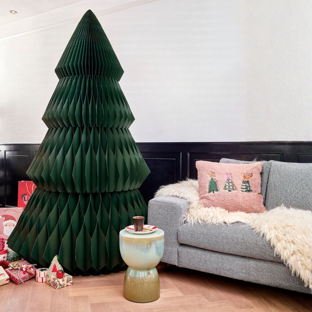 Un sapin de Noël en papier vert est à côté d'un canapé gris avec un coussin rose. Des cadeaux et une petite table avec une tasse sont au premier plan.