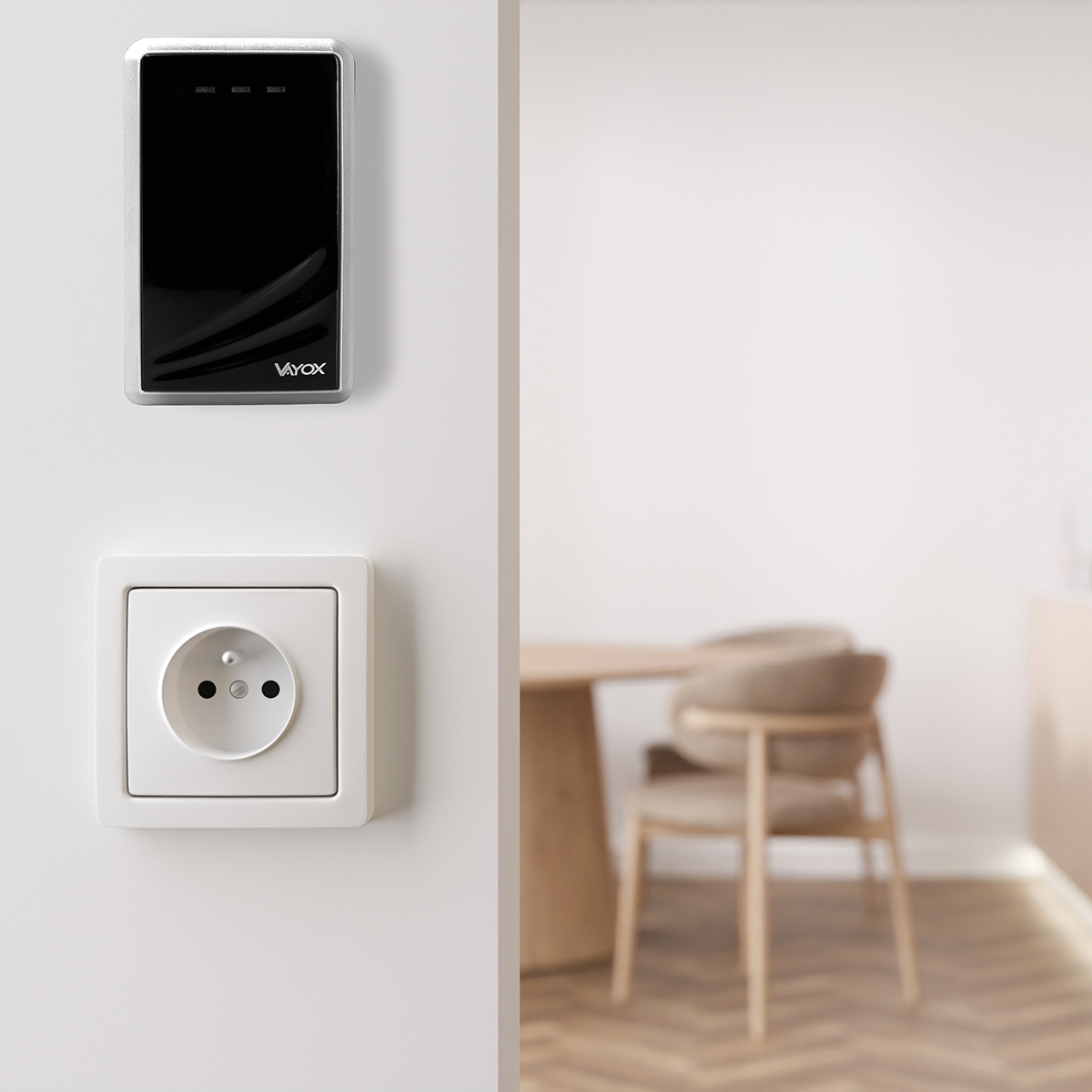 Biała ściana z czarno-srebrnym urządzeniem i białym gniazdkiem. W tle stół i krzesła.