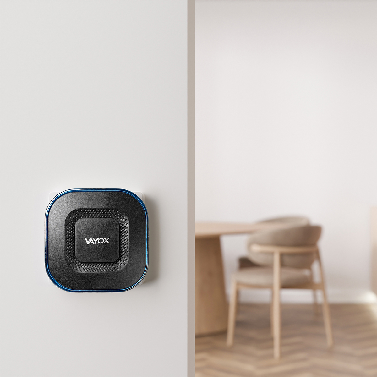 Kwadratowe czarne urządzenie z niebieską ramką jest zamontowane na białej ścianie. Urządzenie ma napis 'Vayox'. Rozmyty zestaw jadalniany jest w tle.