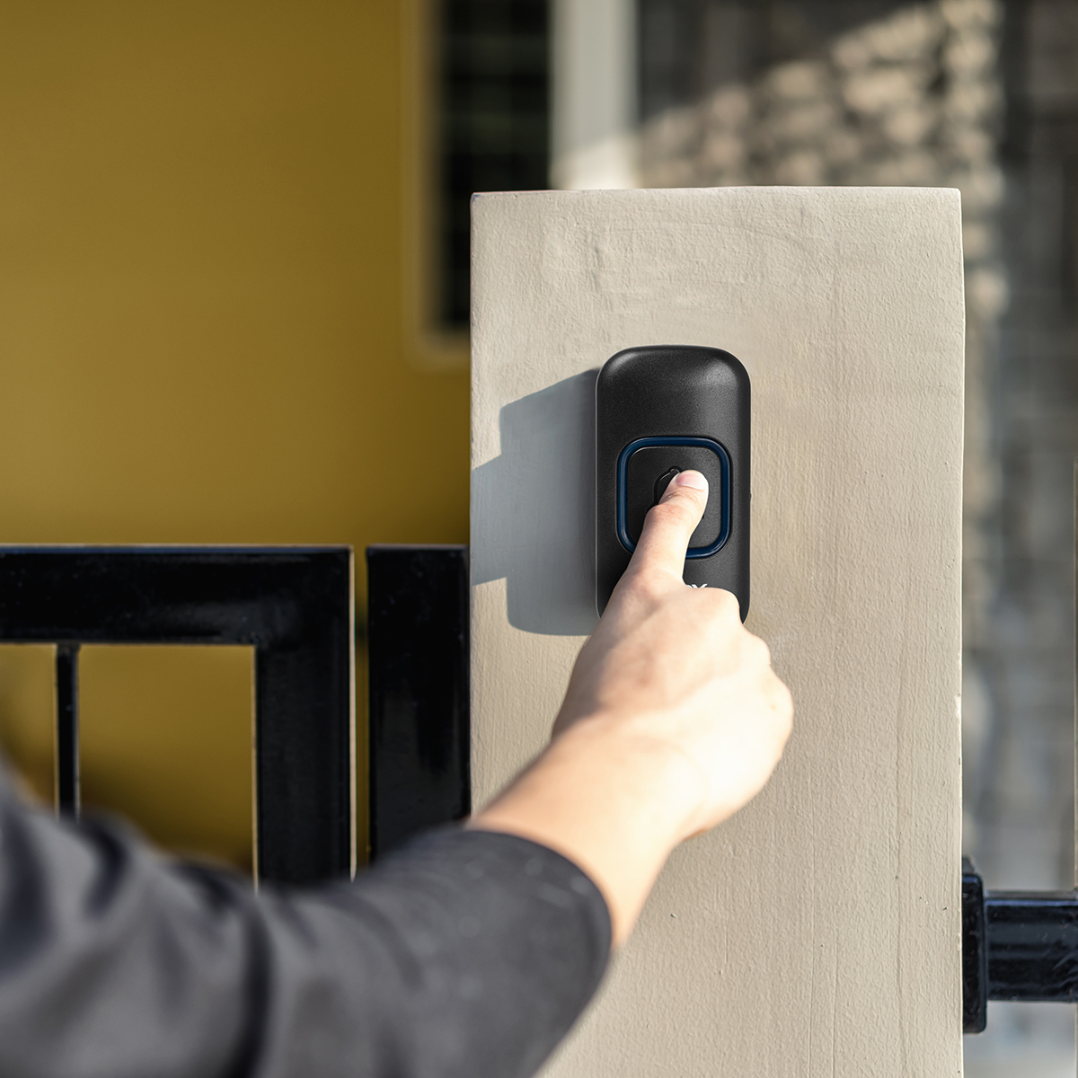 Dłoń naciska dzwonek do drzwi na filarze. Dzwonek jest czarny z niebieskim światłem. W tle czarna brama i żółta ściana.