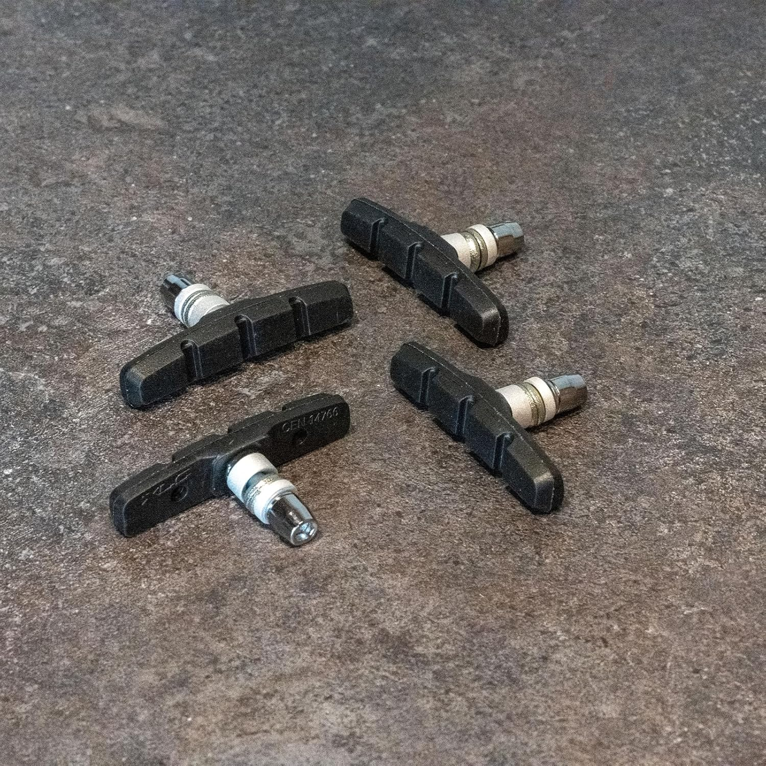 Vier schwarze Fahrradbremsbeläge sind auf einer strukturierten grauen Oberfläche angeordnet. Jeder Belag hat einen silbernen Metallpfosten.