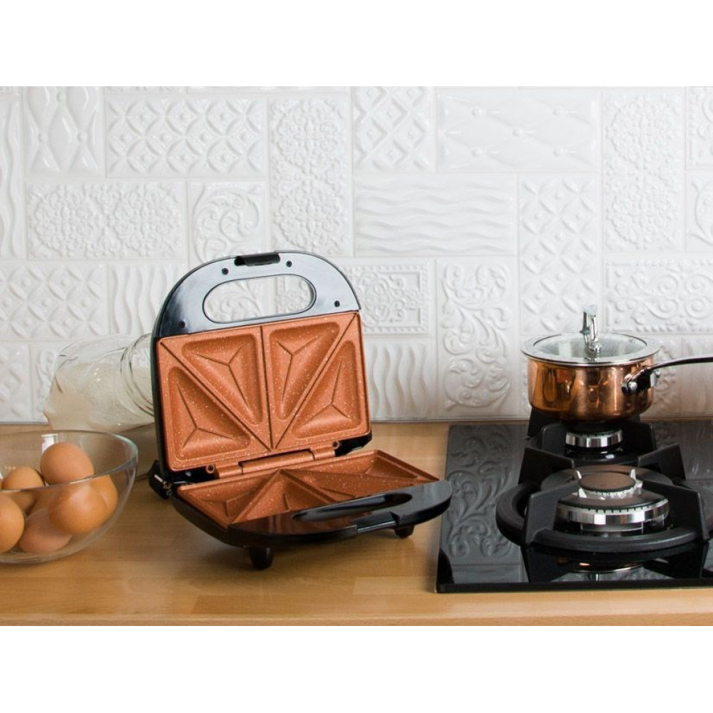 Scena kuchenna z otwartym opiekaczem do kanapek, garnkiem na kuchence, miską jaj i słoikiem. Biała, wzorzysta ściana.