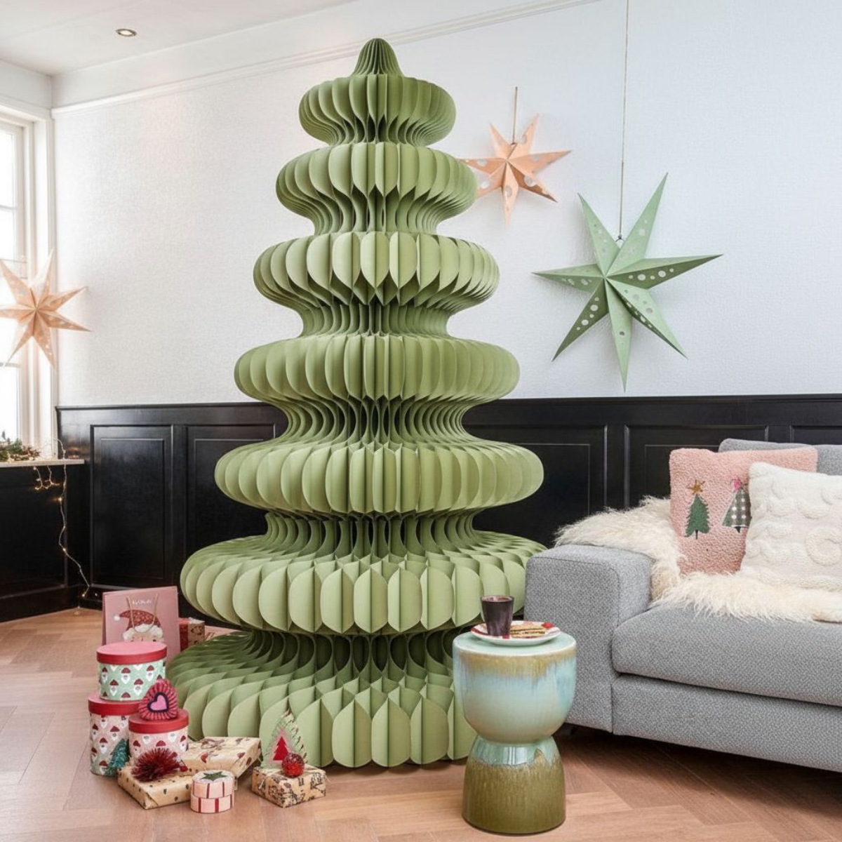 Un sapin de Noël en papier vert est le point central. Des cadeaux sont à la base avec des décorations, des étoiles sur le mur et un canapé gris.