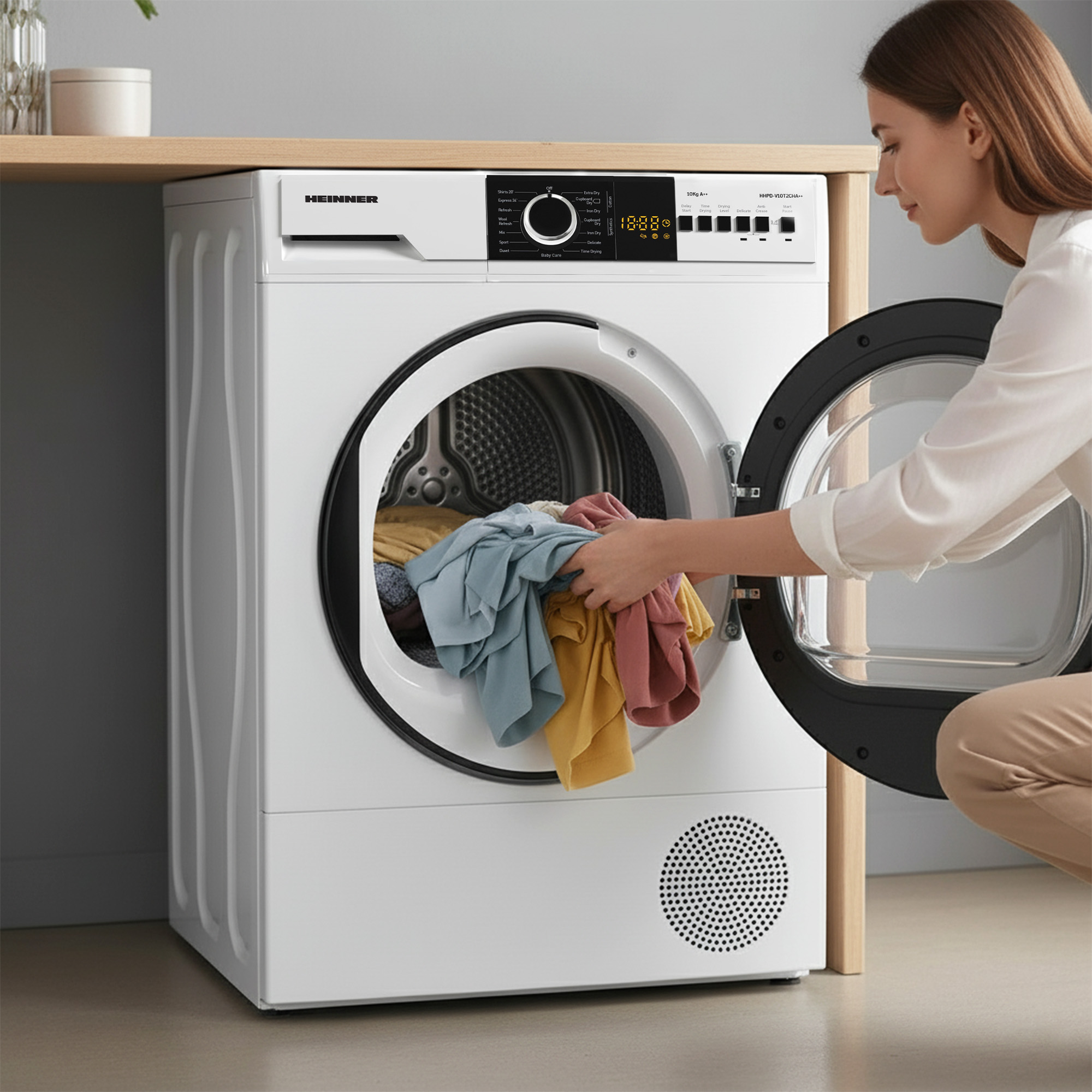 Eine Frau legt Kleidung in einen weißen Trockner. Der Trockner hat ein digitales Display und den Markennamen HEINNER.