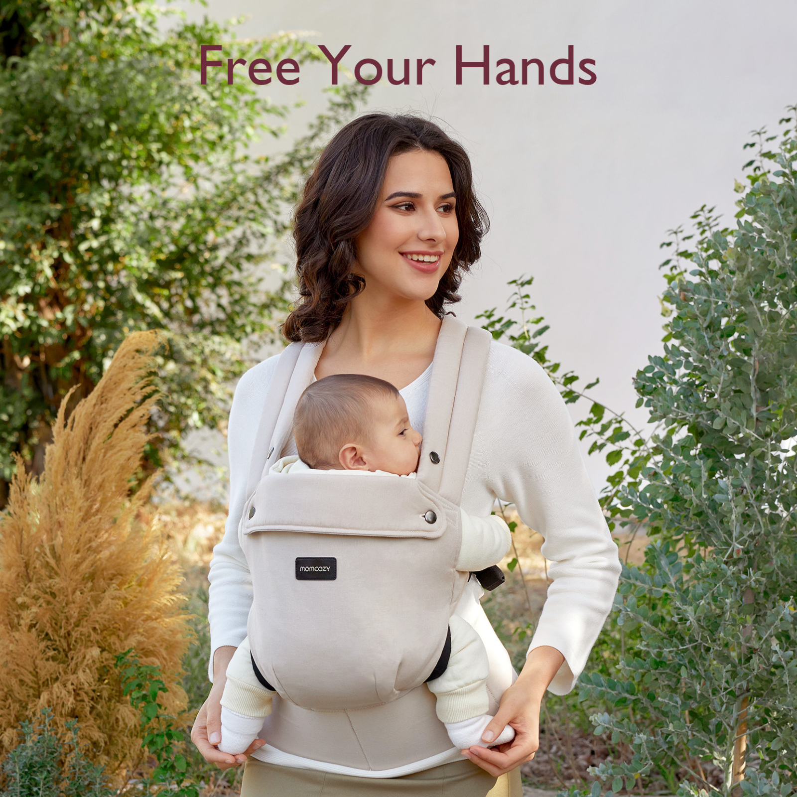 A woman holds a baby in a carrier. They are outdoors. Green bushes in the background. The text says 'Free Your Hands'.